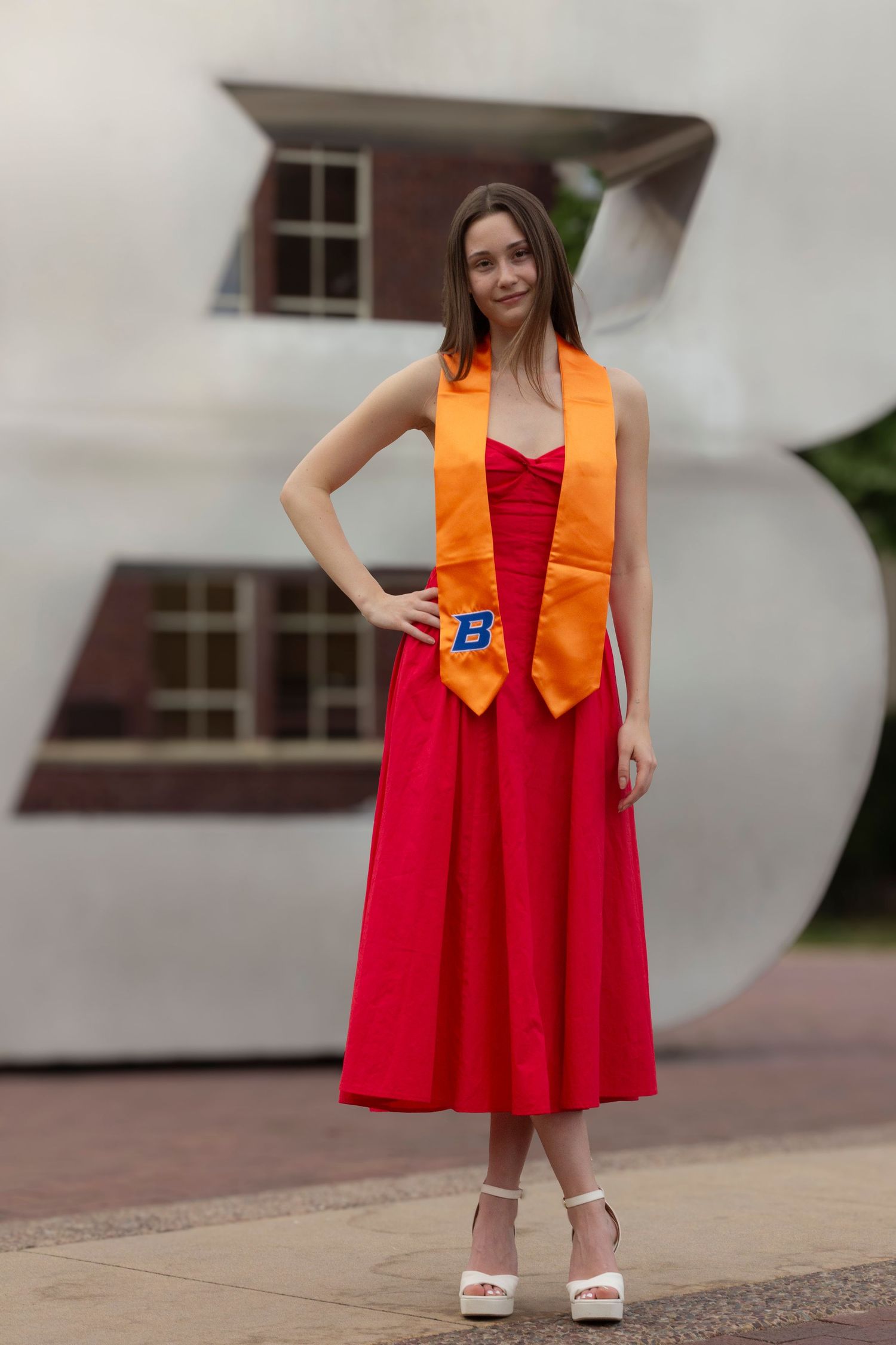 A graduate in a red dress and orange stole with a blue B poses with one hand on her hip in front of a large sculpture.