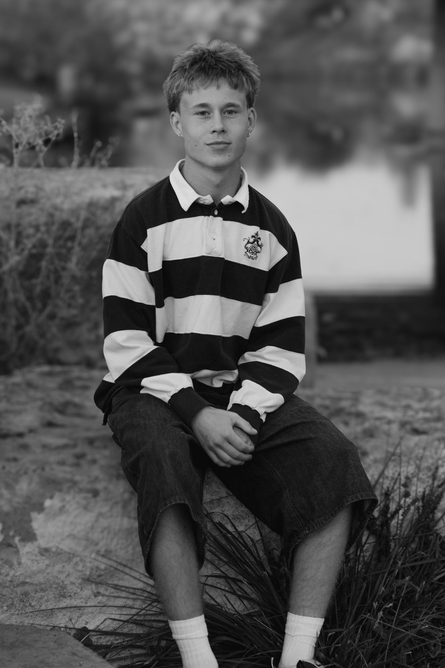 Black and white portrait of a smiling teenage boy in a striped rugby shirt sitting on a rock near the water.