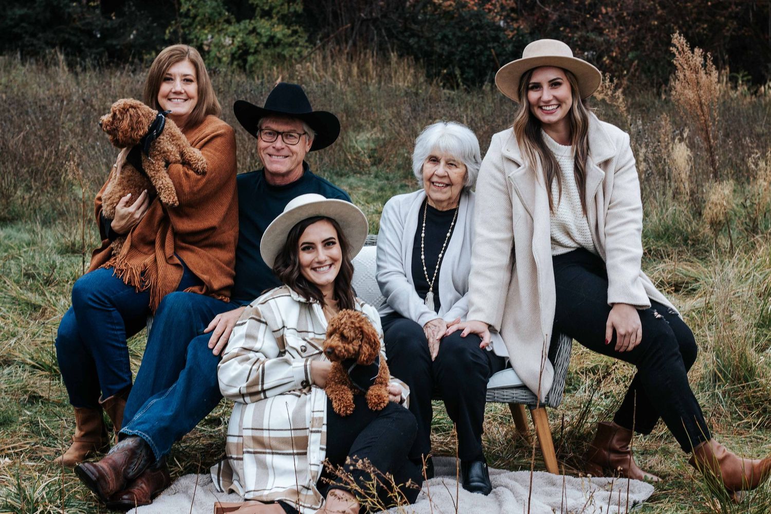 Four women and a man in hats and cozy fall layers sit together outdoors in a field, holding two fluffy puppies.