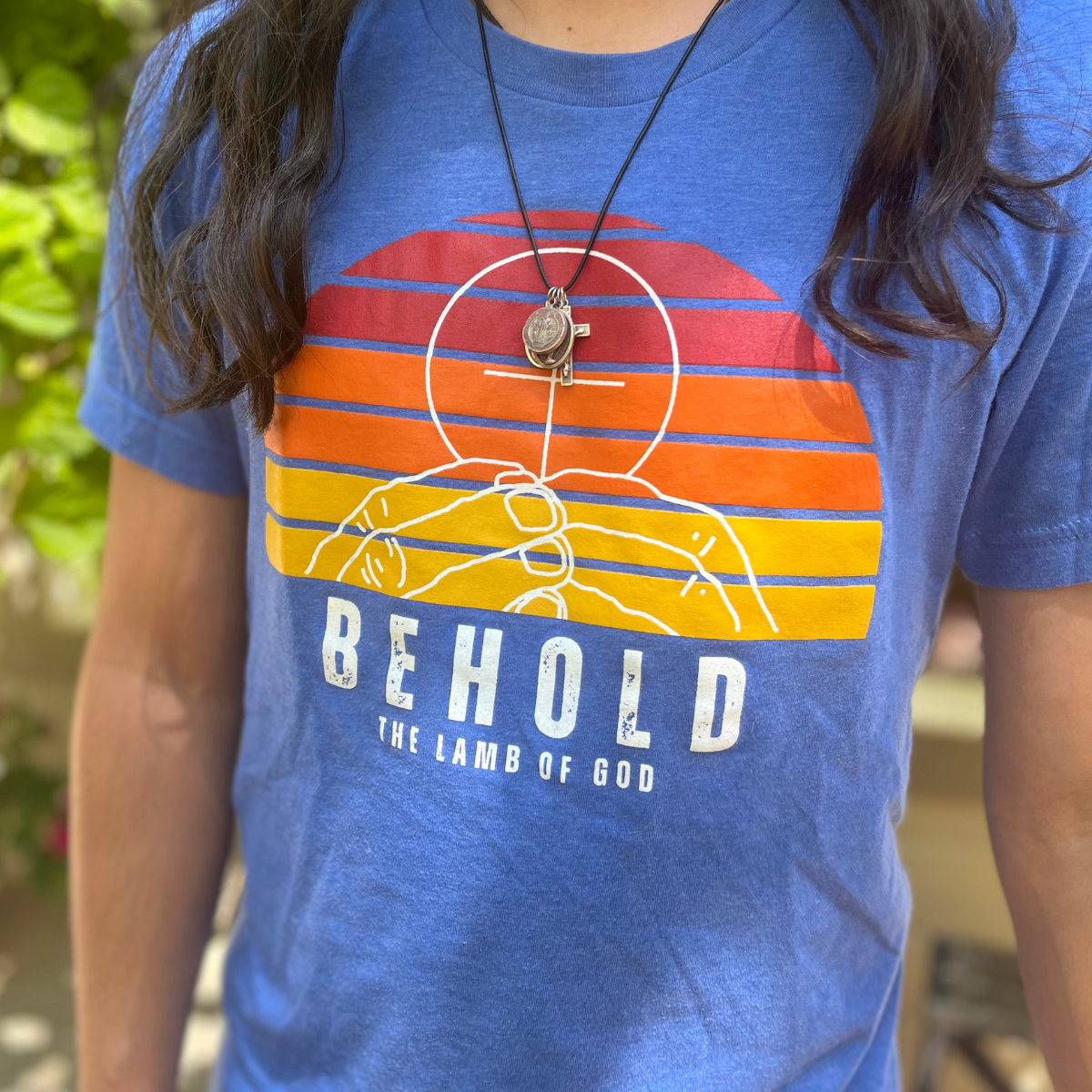 Person wearing a blue t-shirt with a sunset graphic and the words Behold the Lamb of God, plus a pendant necklace.