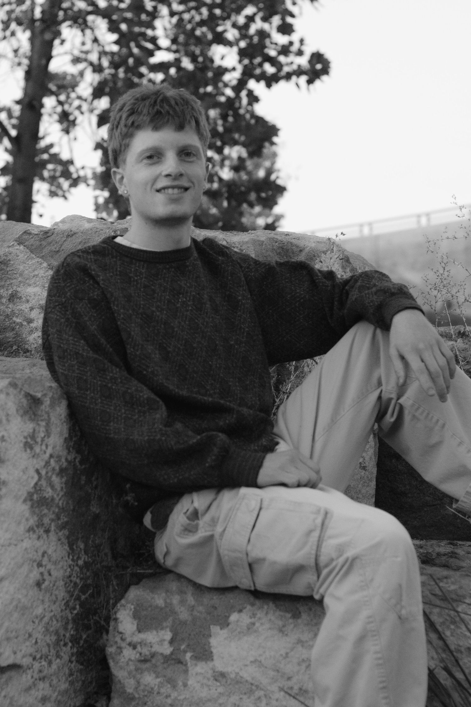 Black and white portrait of a smiling young man in a textured sweater and cargo pants sitting on a large rock outdoors.