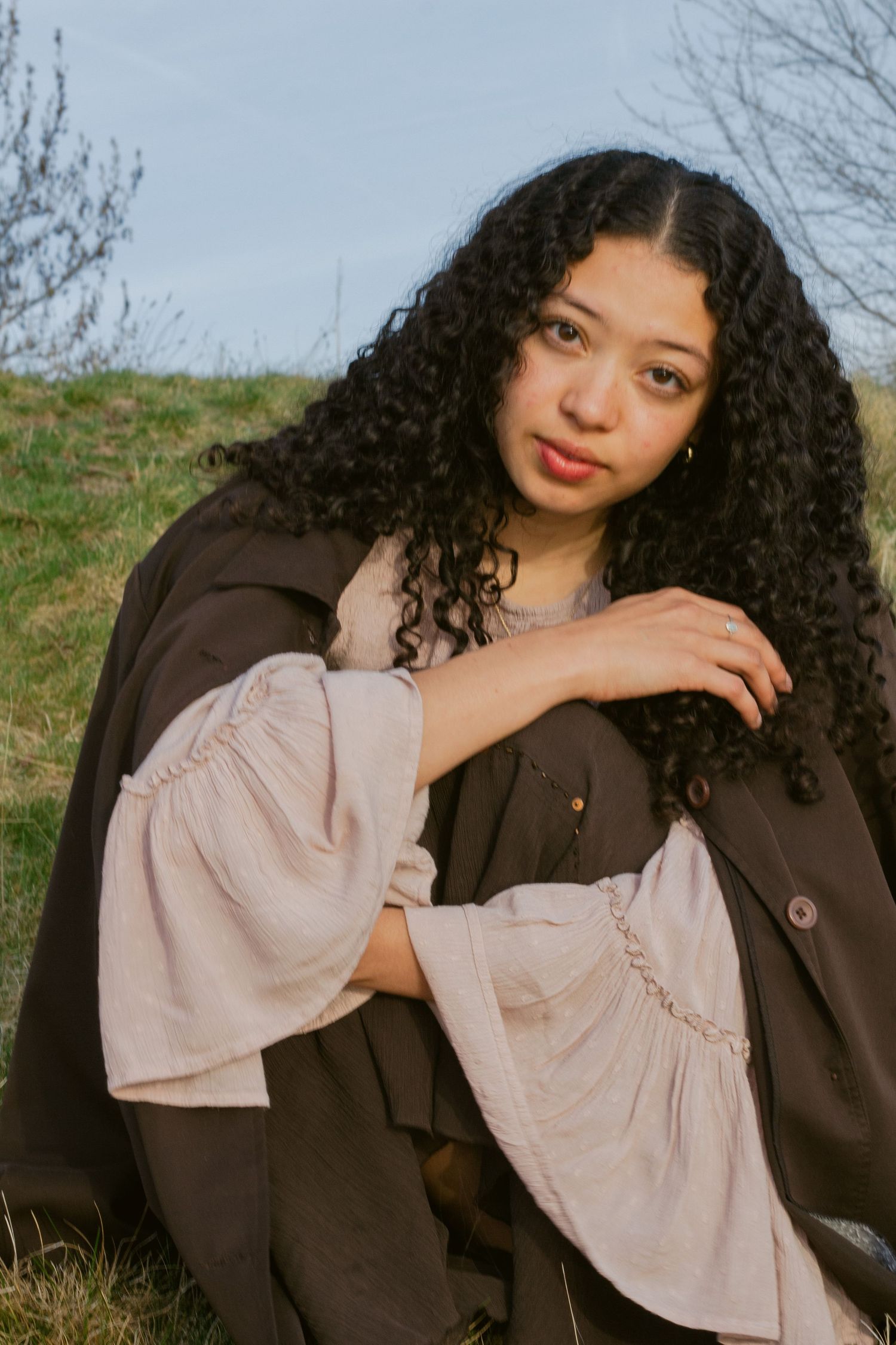 Young woman with long dark curly hair sits on a grassy hillside wearing a brown coat and a blush ruffled-sleeve top.