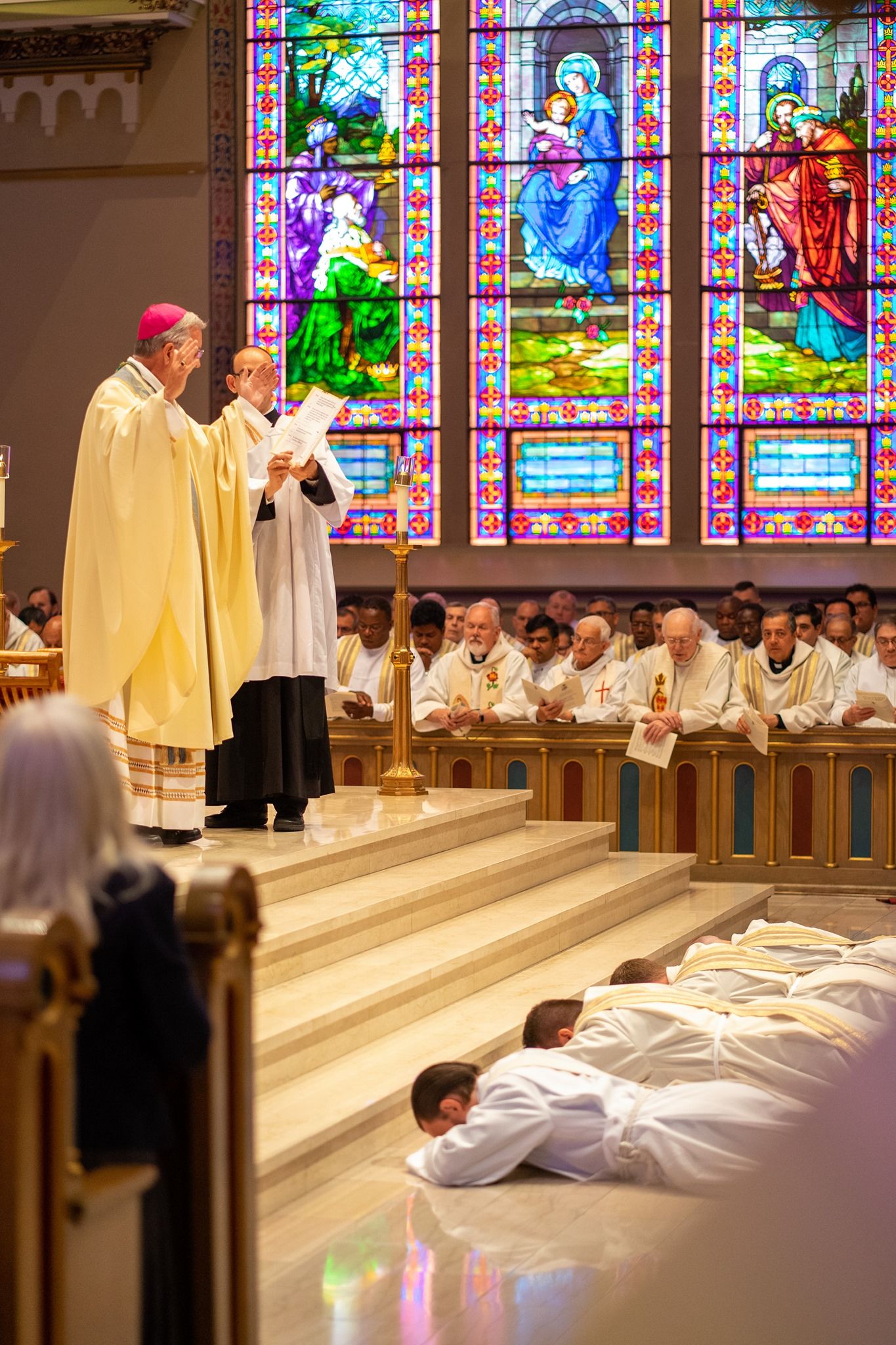 A bishop in a pink zucchetto and gold vestments stands before rows of clergy as ordination candidates lie prostrate on the sanctuary floor.