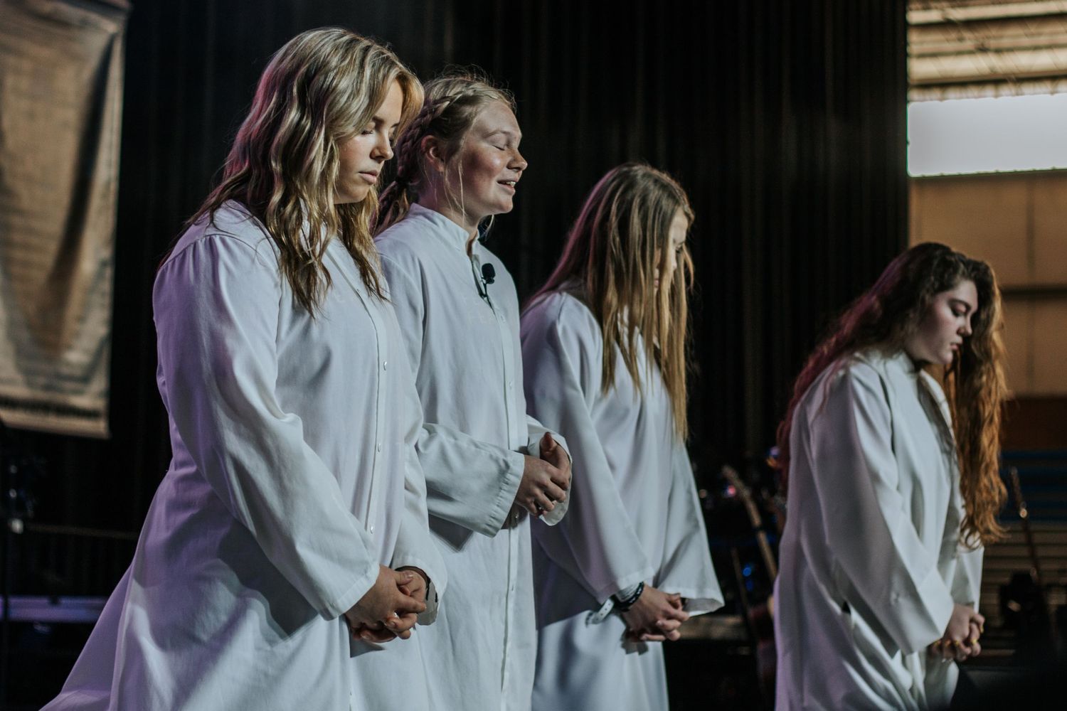 Four young women in white robes stand on stage with hands clasped and heads bowed in a reverent moment of prayer.