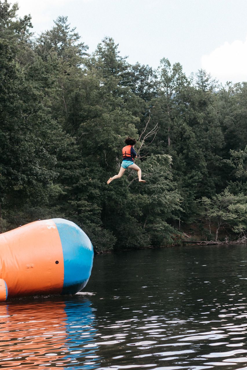 A person in an orange life vest leaps through the air toward an inflatable water blob floating on a forest-lined lake.