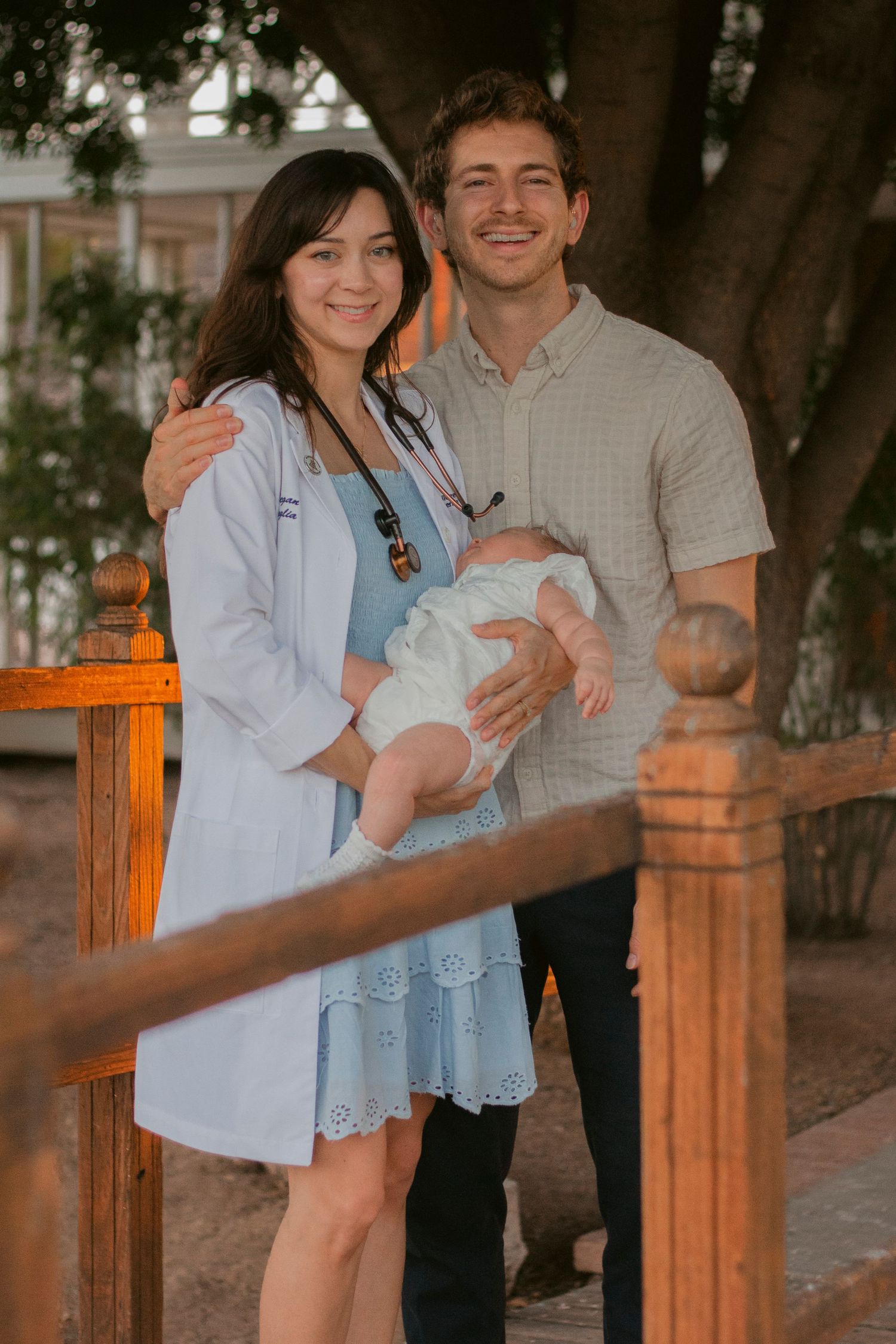 Smiling couple on a wooden porch hold their newborn baby, the mother wearing a white coat with a stethoscope over a blue dress.