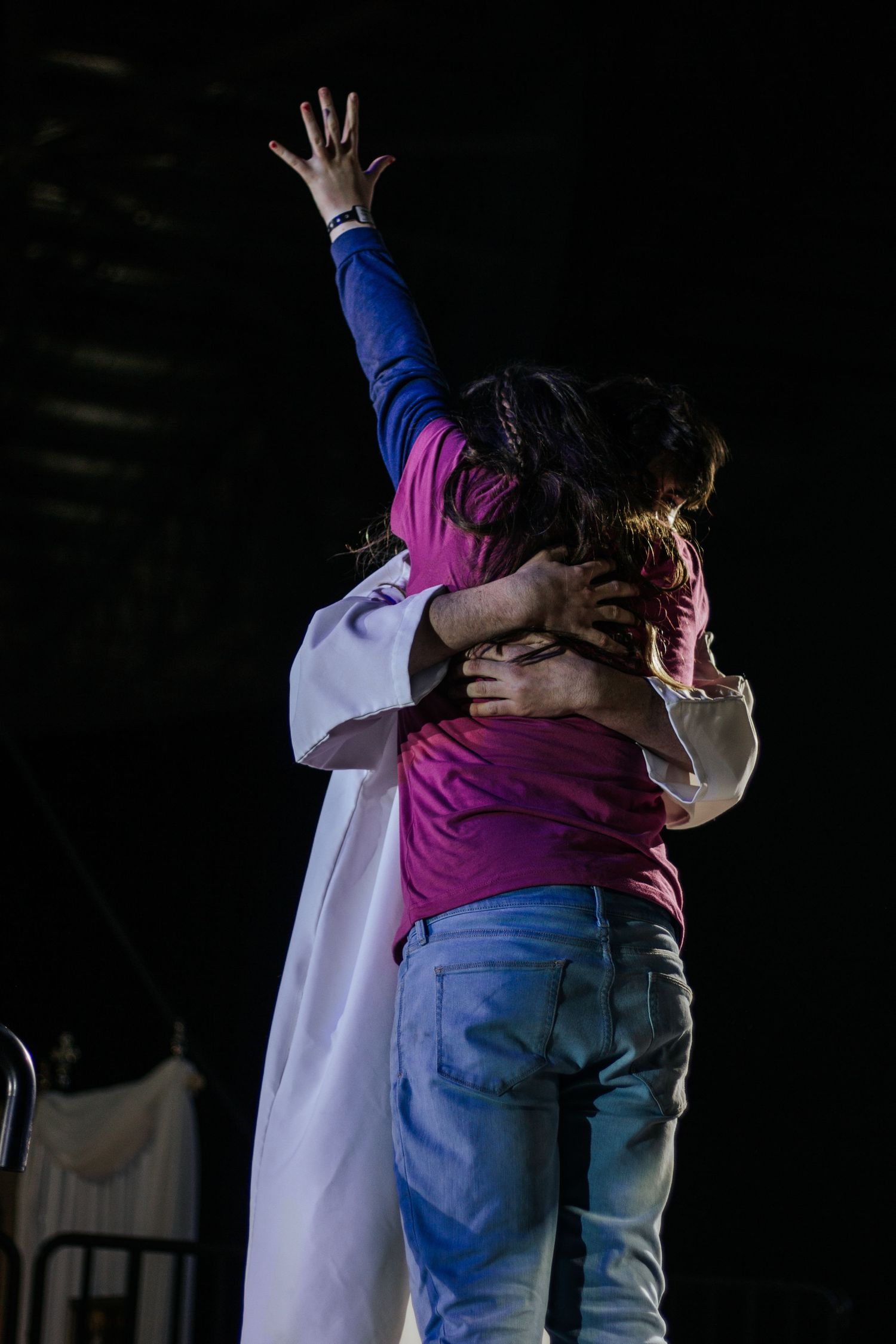 Person in a pink shirt raises one hand while embracing someone in a white robe on a dark stage in a moving performance.