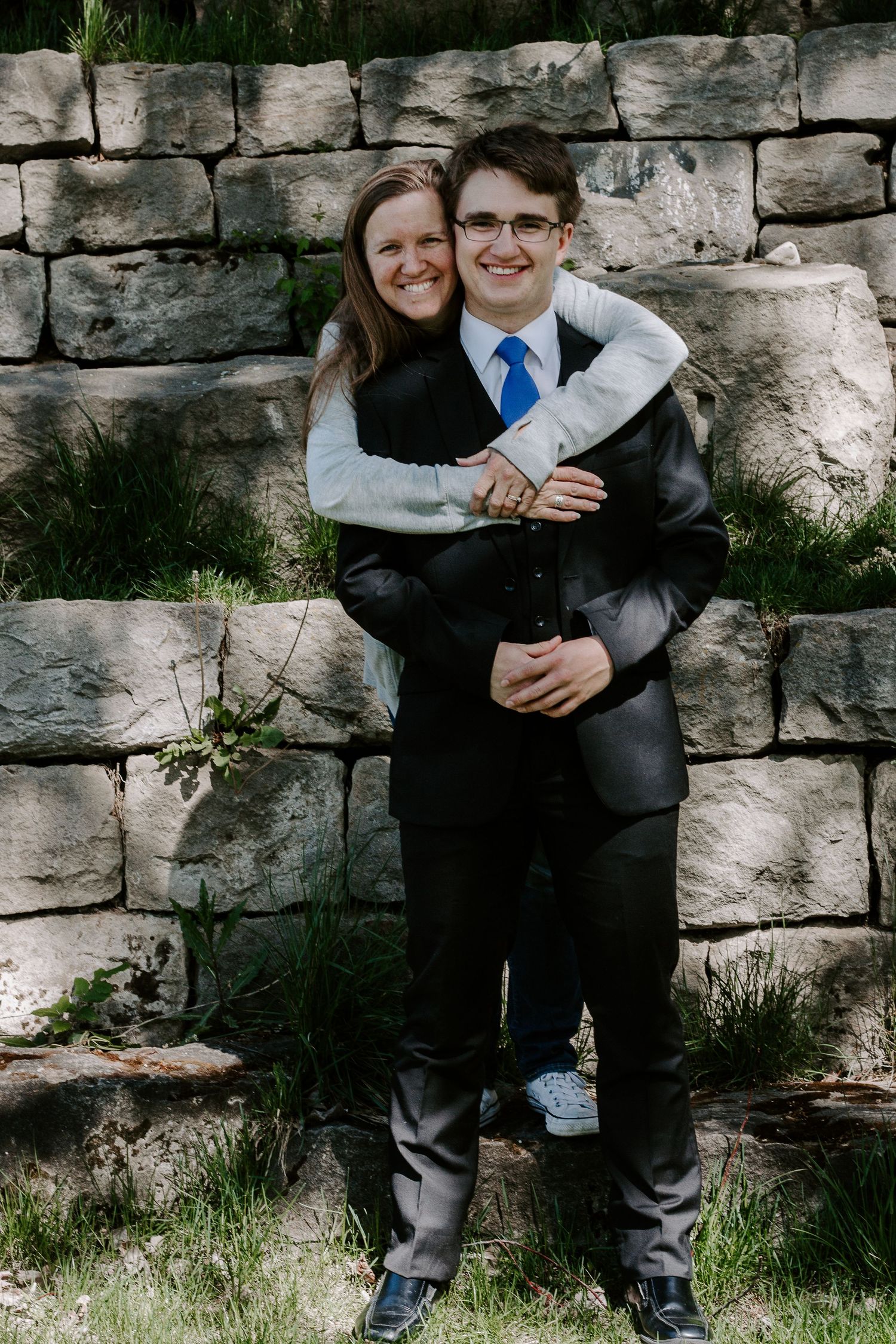 Young man in a suit and blue tie smiles while a woman hugs him from behind in front of a stone wall.