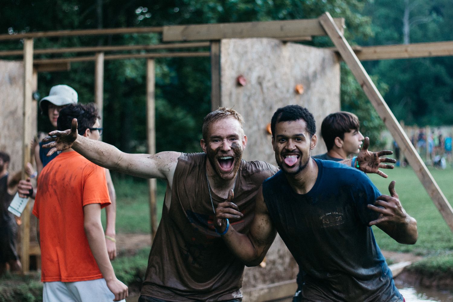 Two muddy, laughing men pose with arms outstretched near a wooden climbing wall at an outdoor obstacle course.