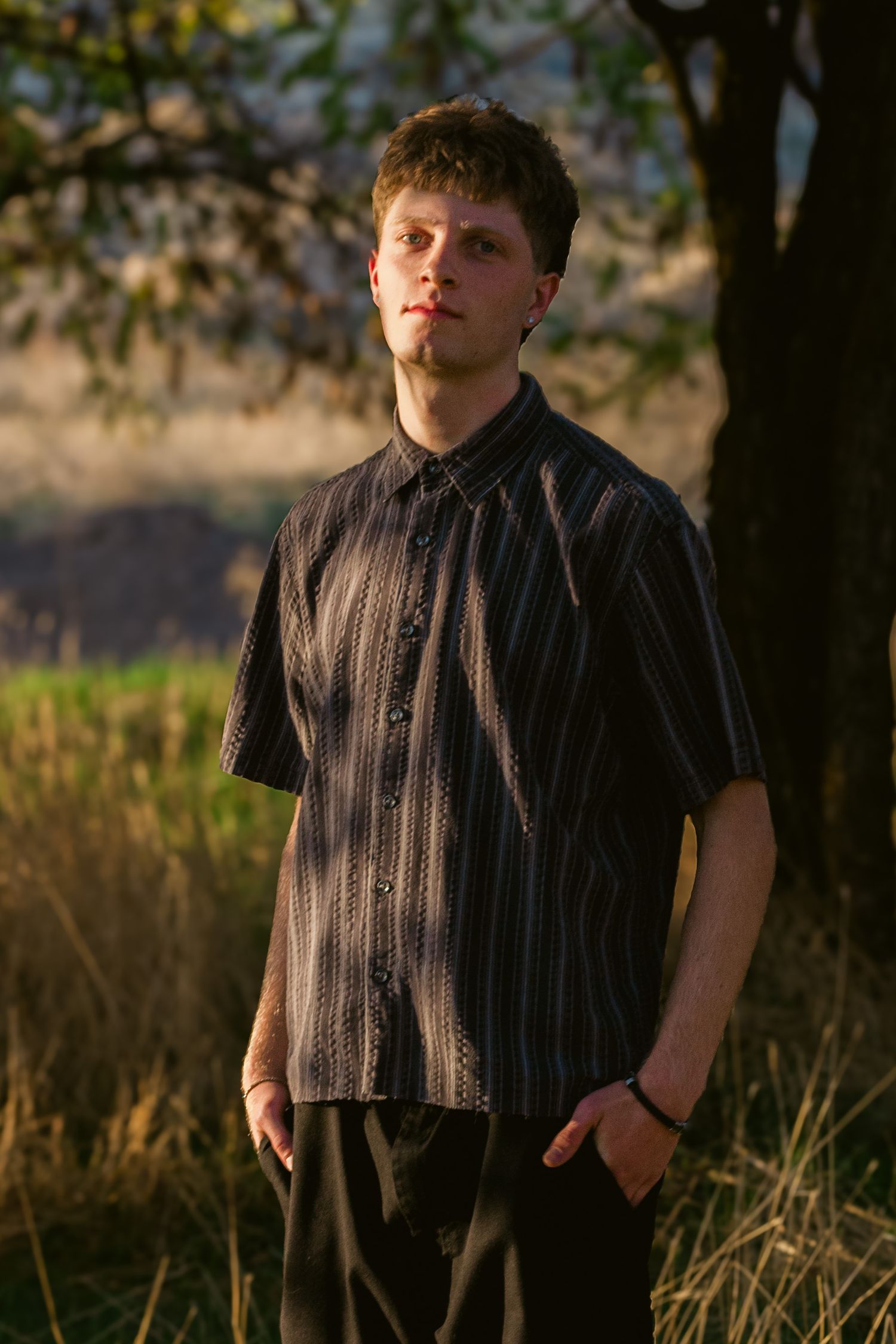 Young man in a dark striped short-sleeve shirt stands with hands in pockets among tall dry grass and trees in warm light.