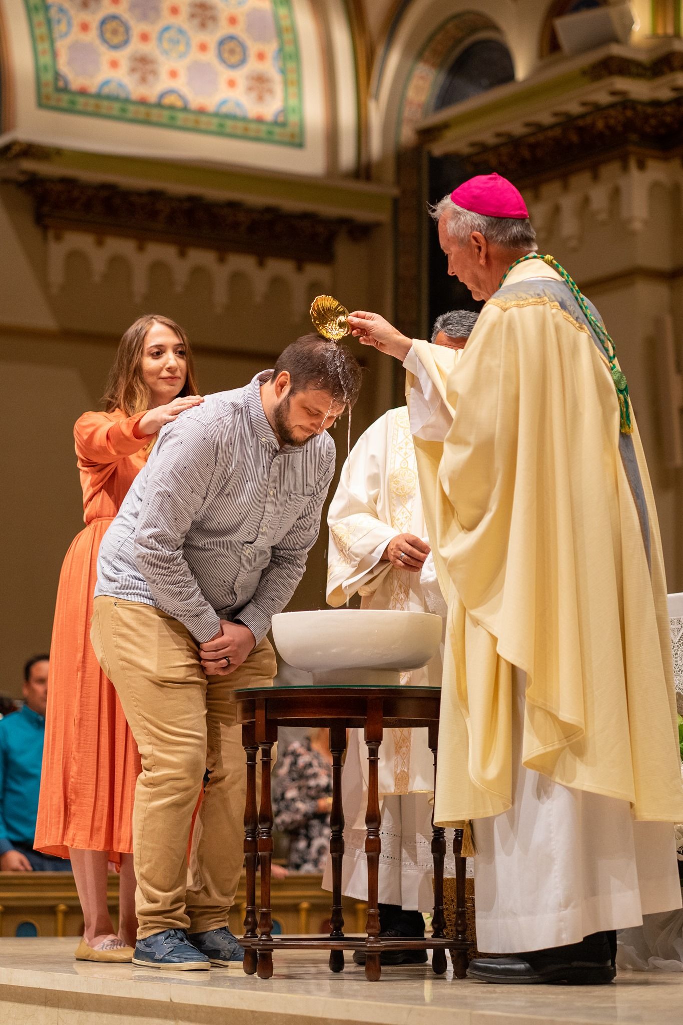 A bishop pours water over the head of a man leaning over a baptismal font while a woman in an orange dress supports him in church.