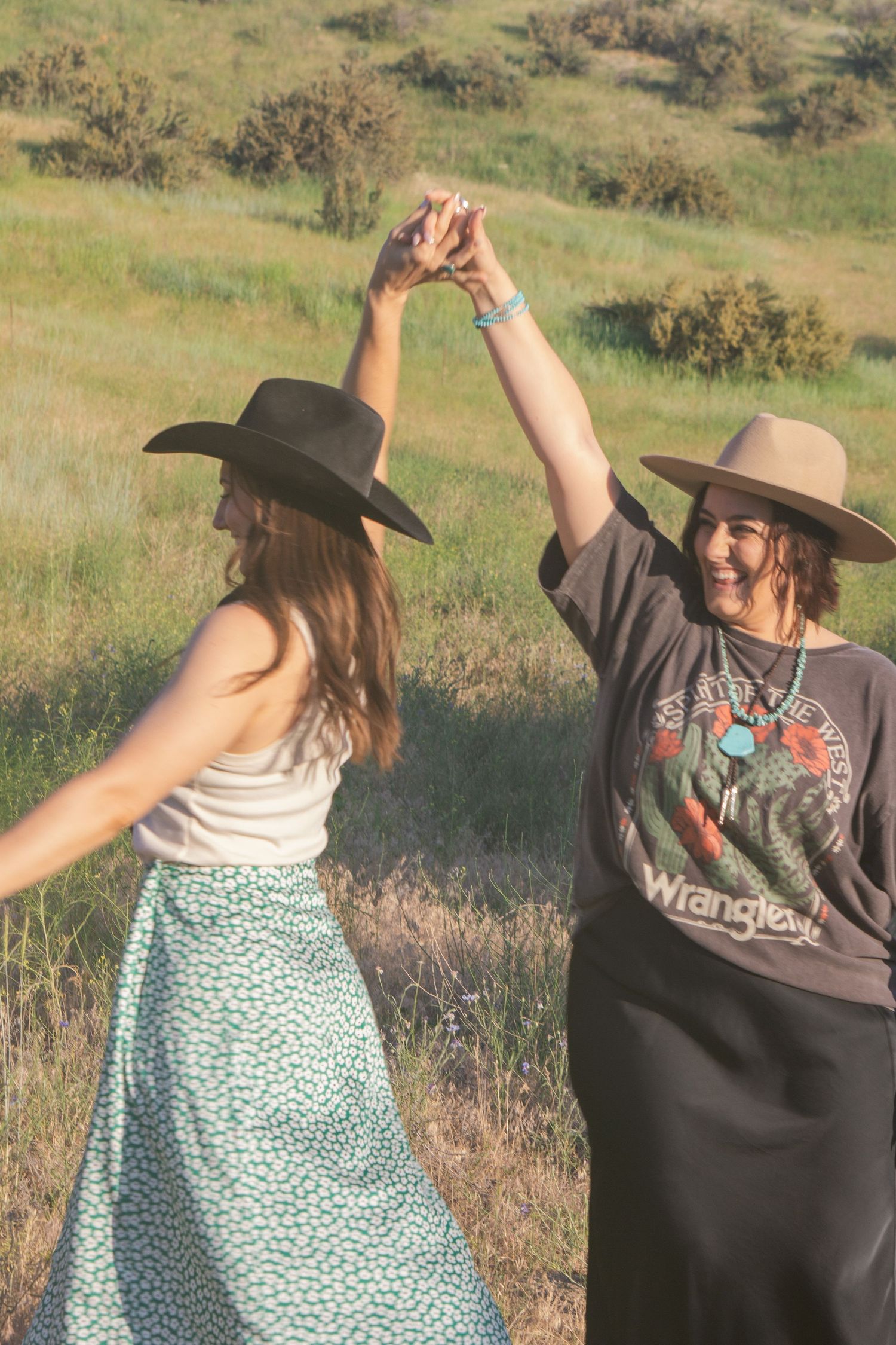 Two joyful women in hats hold hands raised overhead and dance together in a sunny green meadow.