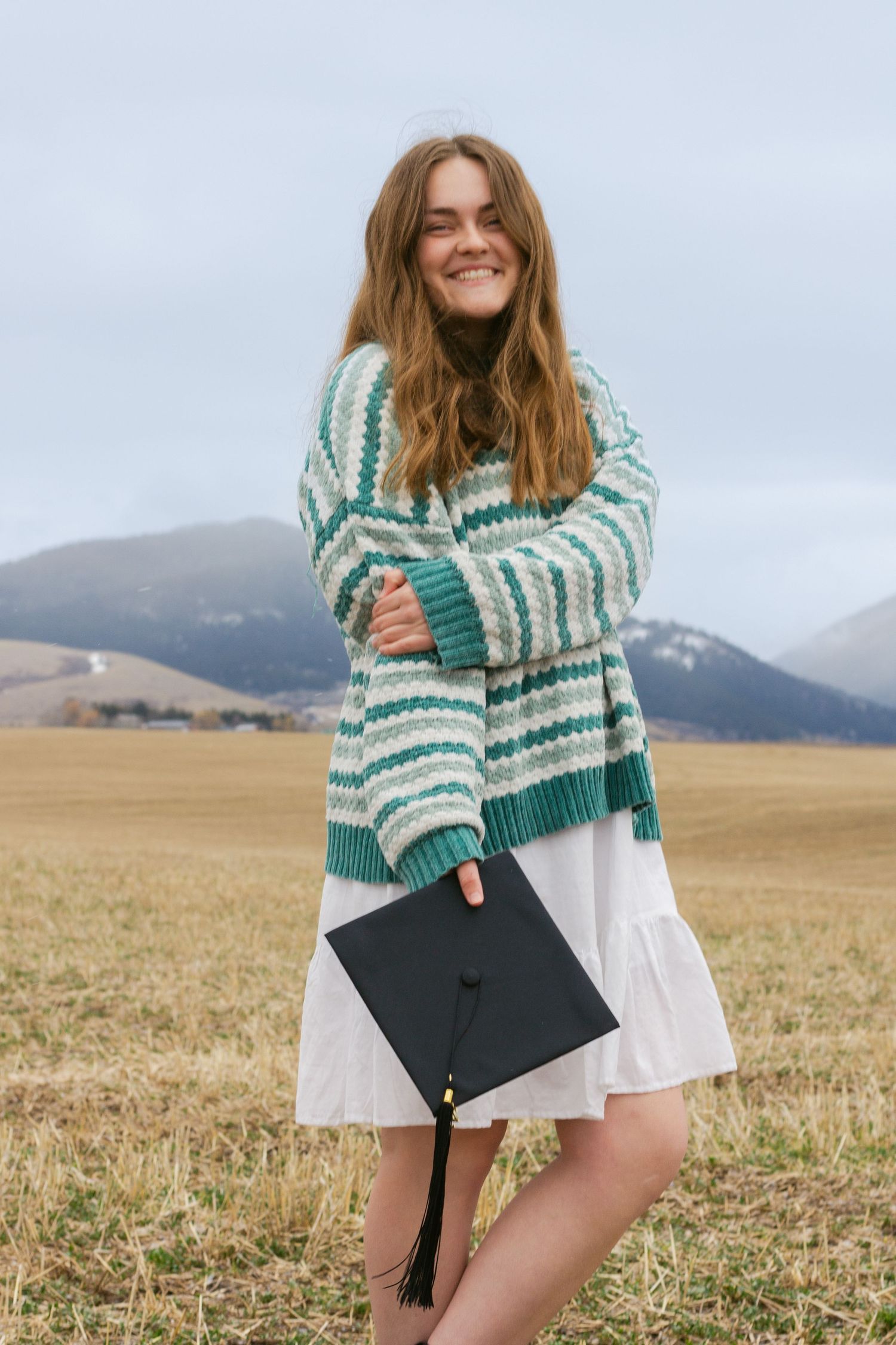 Smiling young woman in a green striped cardigan and white skirt holding a black graduation cap in a field with mountains behind her.