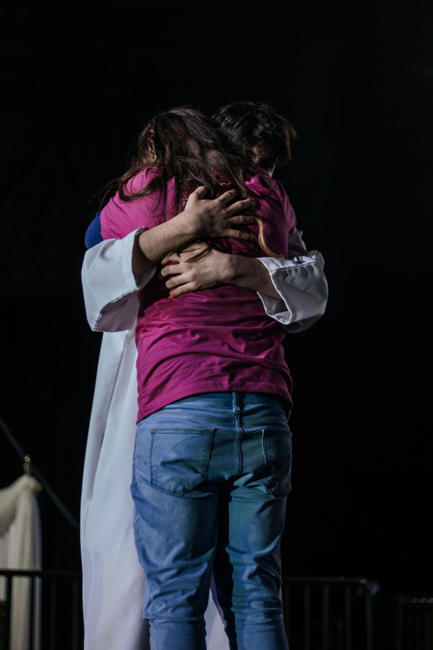 Person in a pink shirt and jeans embraces someone in a white robe on a dark stage in an emotional moment.