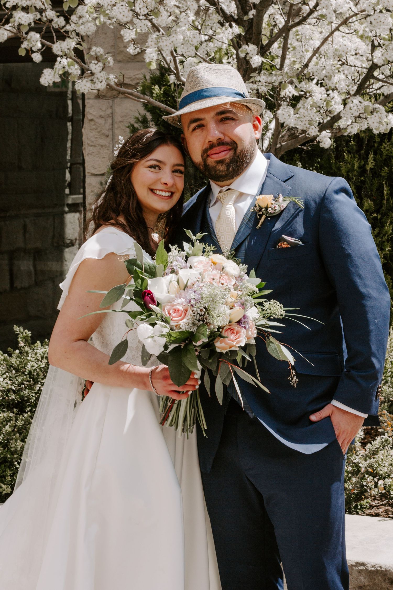 A happy newlywed couple poses outdoors, the bride holding a pink and white bouquet and the groom wearing a navy suit and straw hat.