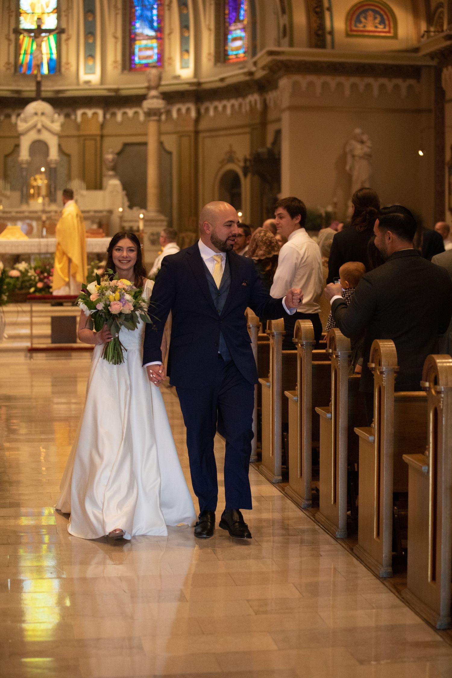 A bride and groom walk hand in hand down the aisle of a catholic church as guests seated in wooden pews look on.