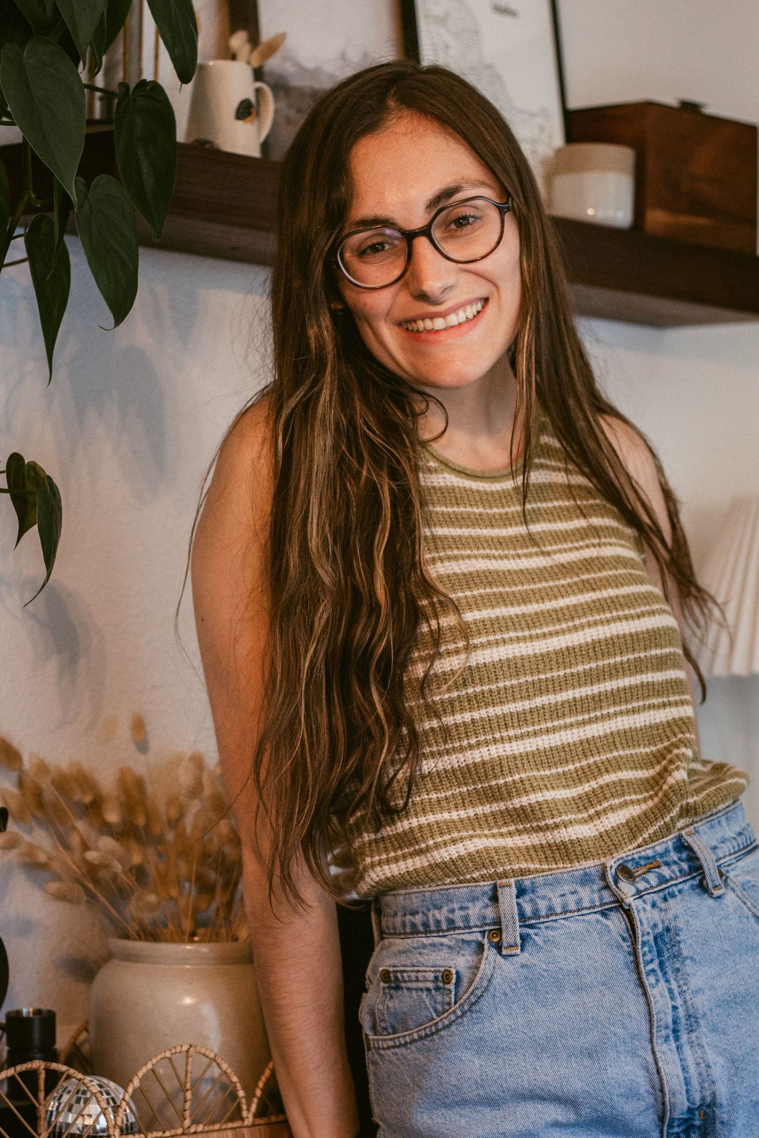 A smiling woman with long brown hair and glasses wears a striped tank top and denim jeans, standing beside dried flowers in a cozy home.