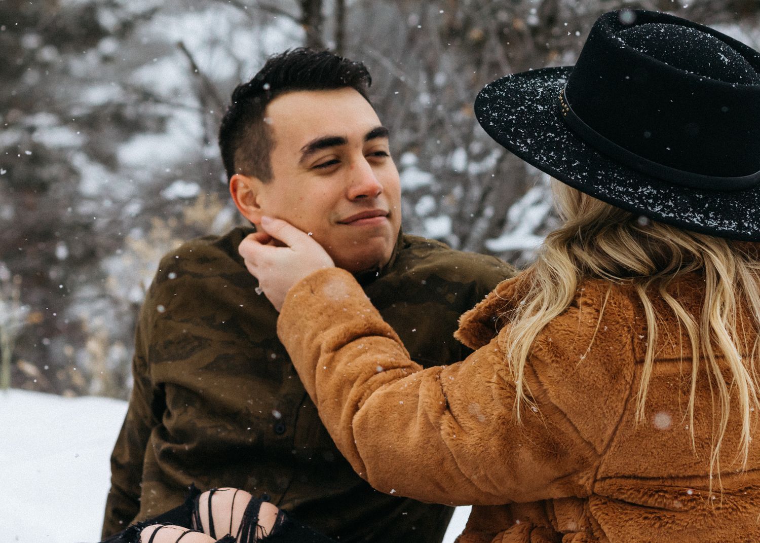 Woman in a black sparkly hat and brown fur coat gently touches her smiling partner's face in falling snow.