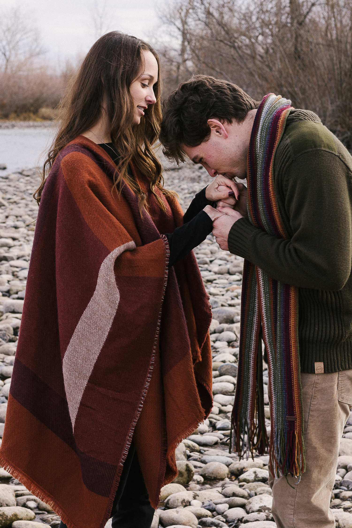 Man in a striped scarf tenderly kisses a woman's hand as she smiles, standing on a pebbly riverbank in a wool wrap.