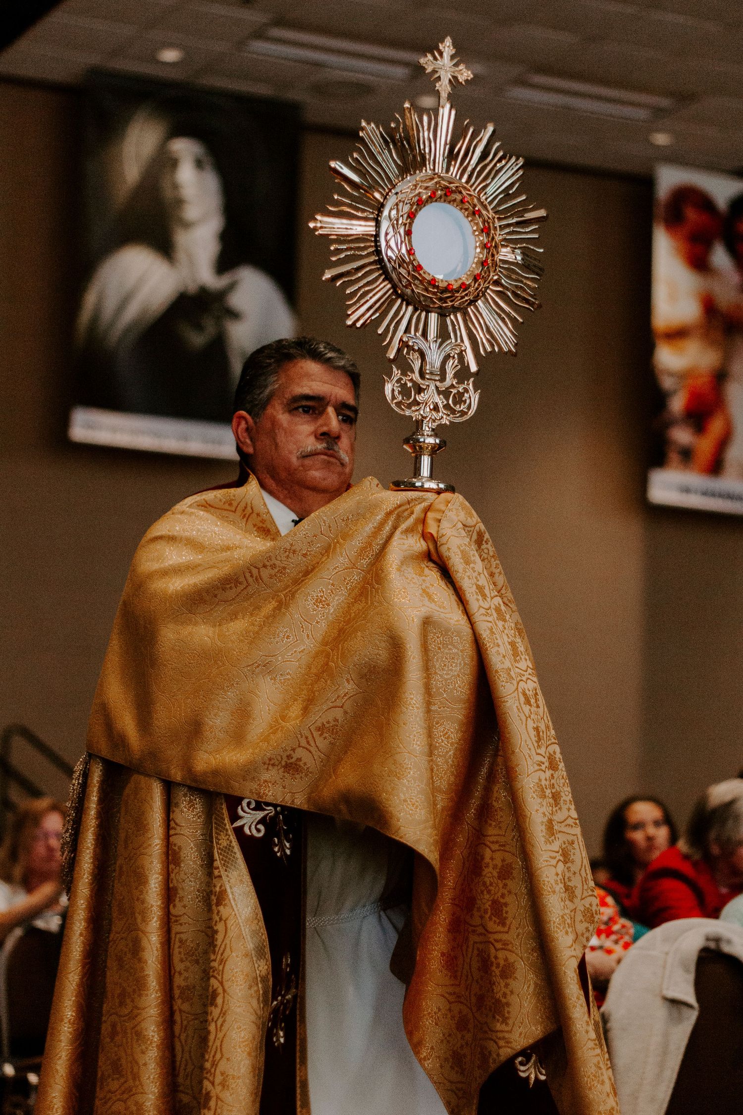 A deacon carries a radiant monstrance during a Eucharistic procession in an indoor hall. Catholic women's conference