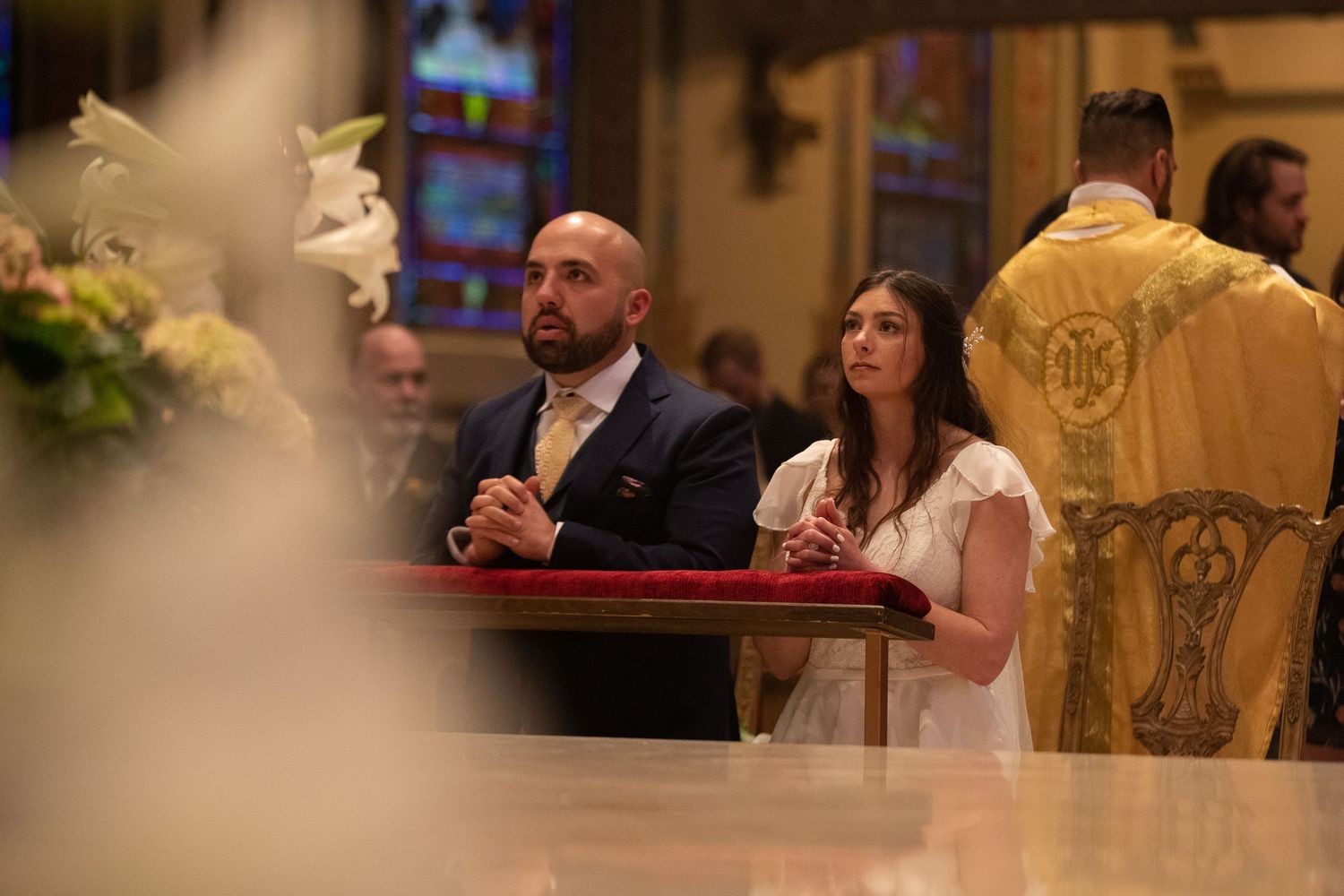 A groom and bride kneel in prayer at a red-draped altar during a church wedding ceremony as a priest in gold vestments stands nearby.