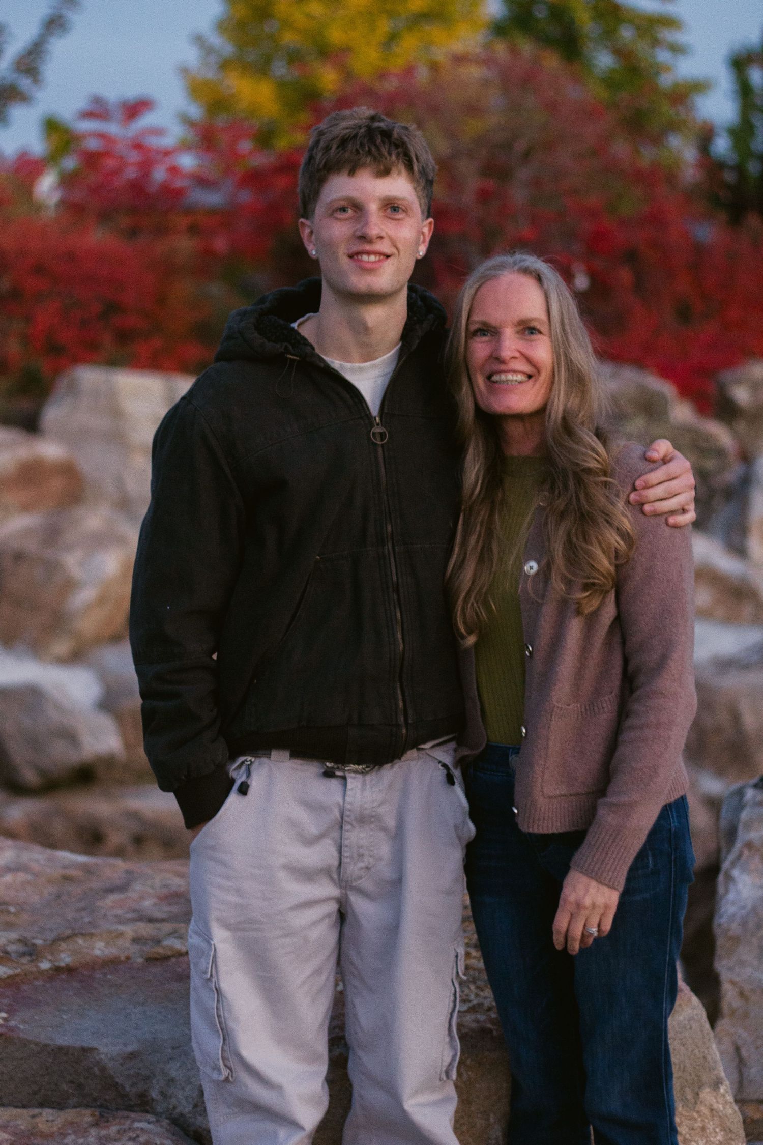 Smiling young man and mother embrace in front of boulders and vibrant red autumn foliage, both dressed casually in jackets and jeans.