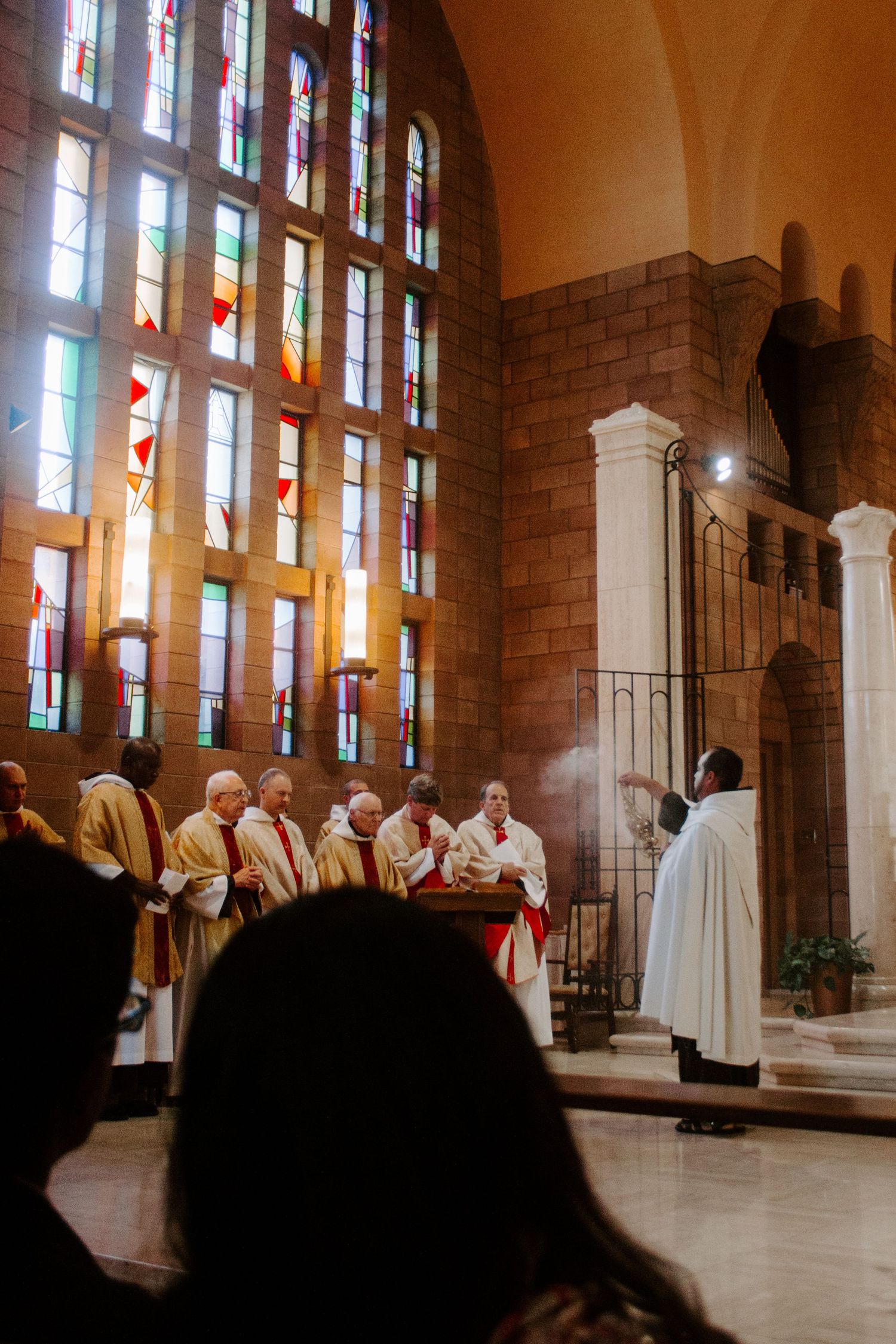 Priests in vestments gather at an altar during Mass in a church with tall stained glass windows while a robed server lifts incense.