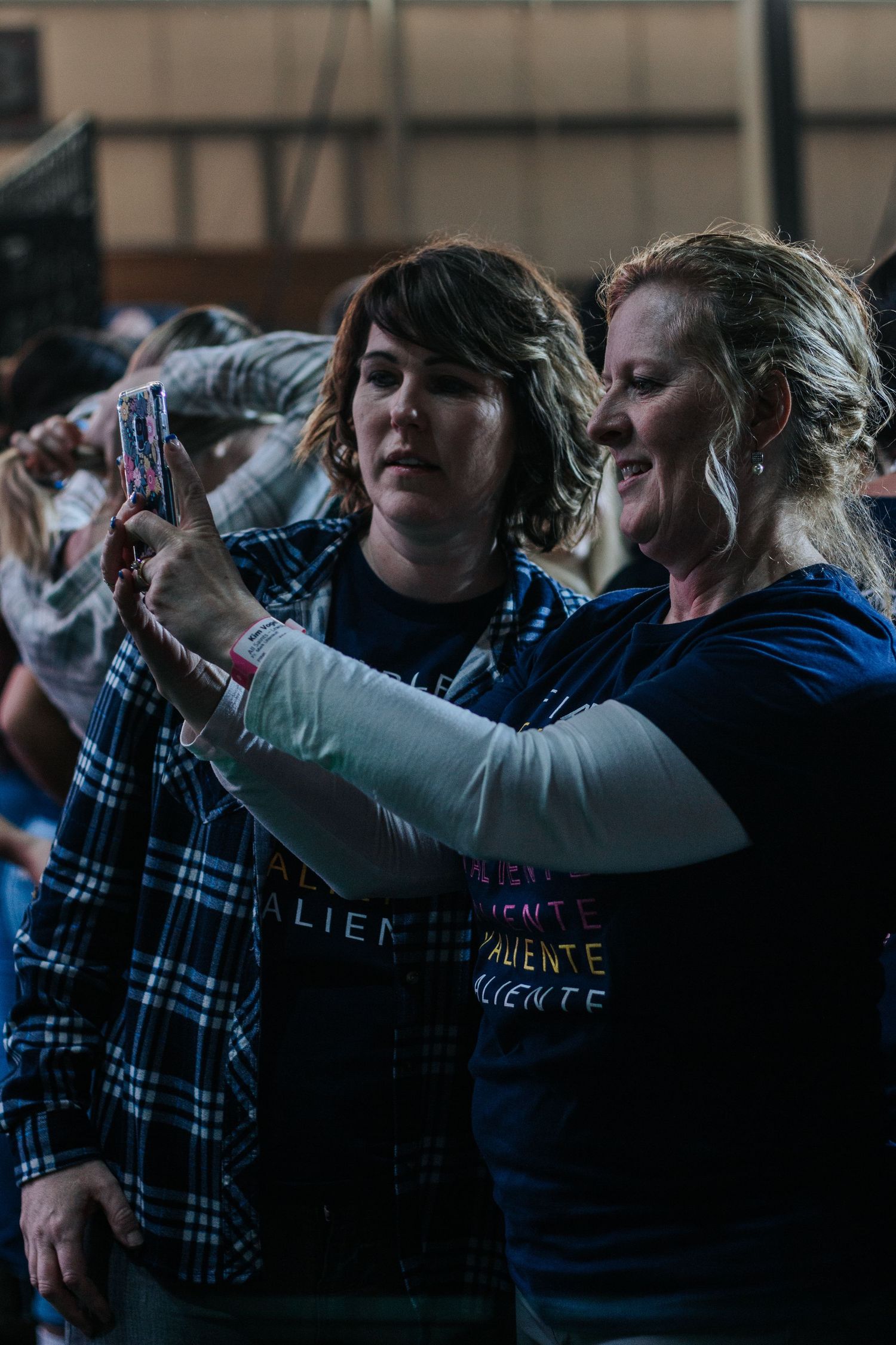 Two smiling women in navy event t-shirts take a selfie with a phone while standing in a crowd at a conference.