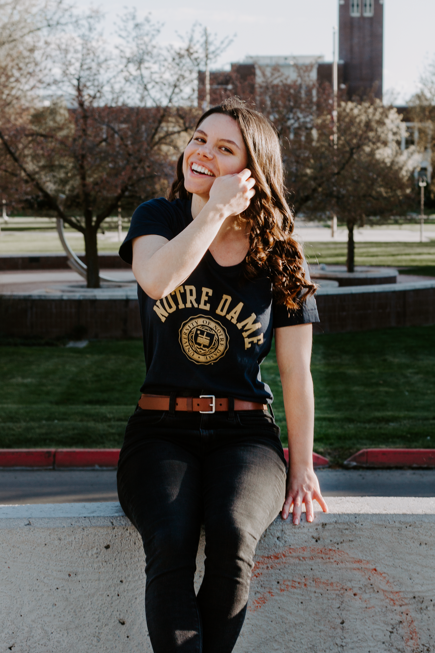 A smiling woman in a Notre Dame t-shirt and brown belt sits on a low wall on a sunny campus lawn.