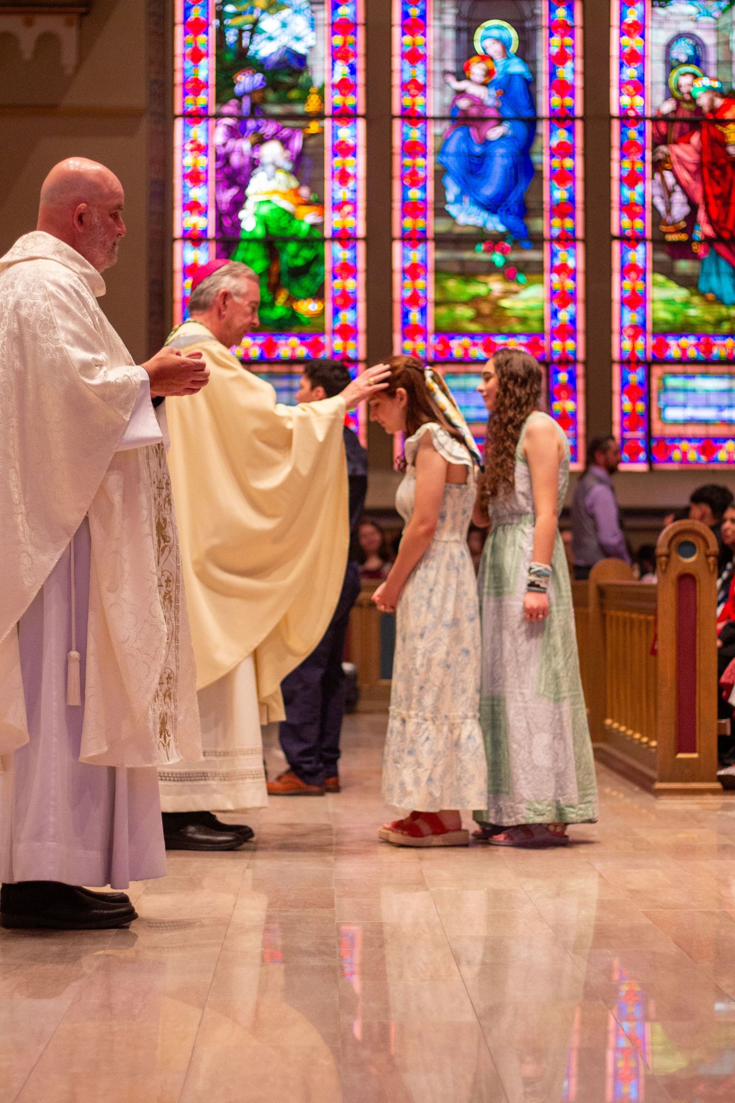 A priest in a cream chasuble blesses two girls in white dresses before stained glass windows during a Catholic church ceremony.