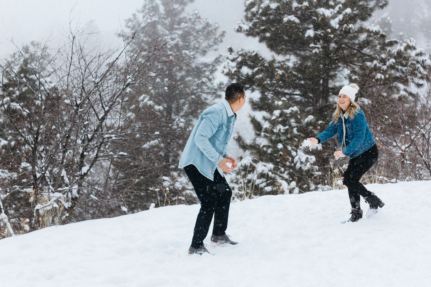 A couple plays in a snowy field, preparing to throw snowballs at each other beside snow-covered evergreen trees.