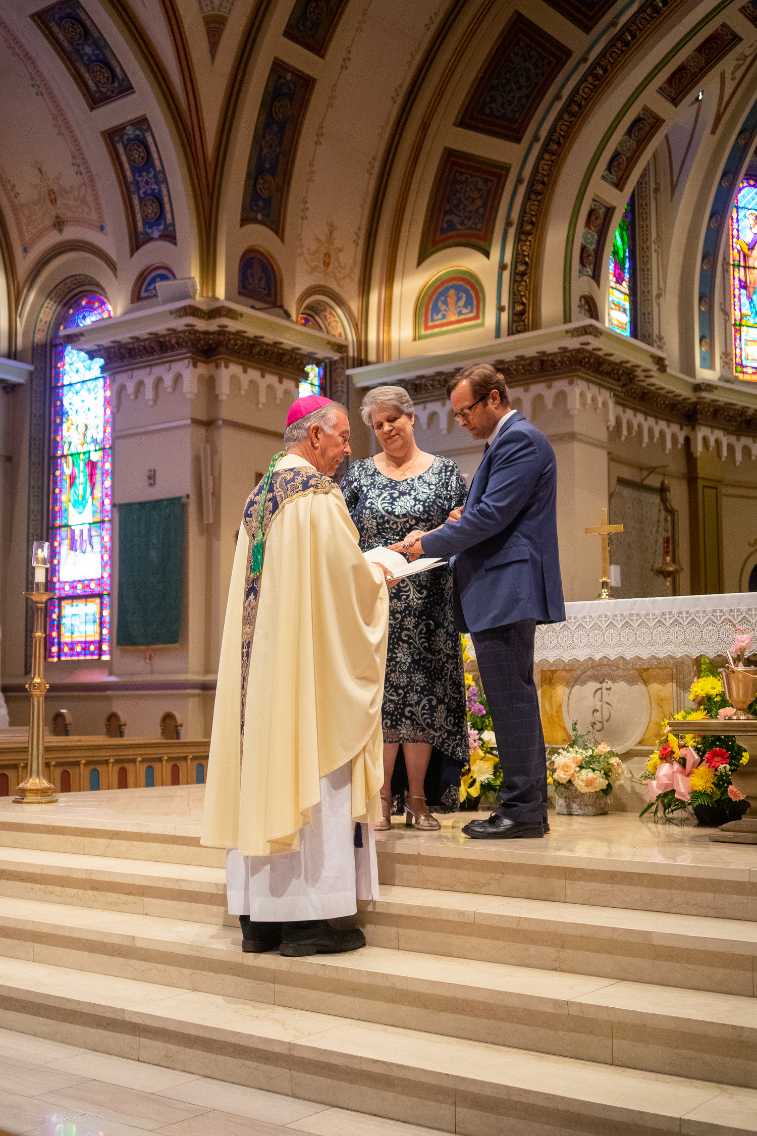 A bishop in a pink zucchetto and vestments reads from a book beside a smiling woman and a man at the church altar.
