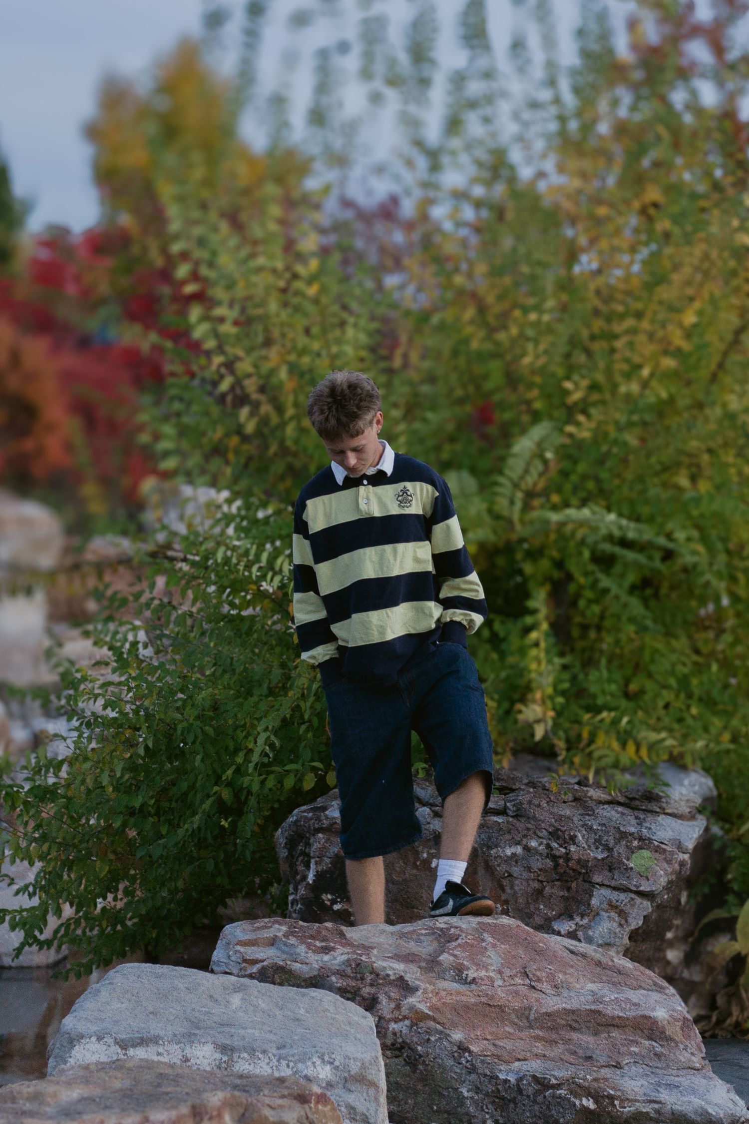 Teenage boy in a striped rugby shirt and dark shorts stands on large rocks surrounded by colorful autumn foliage.