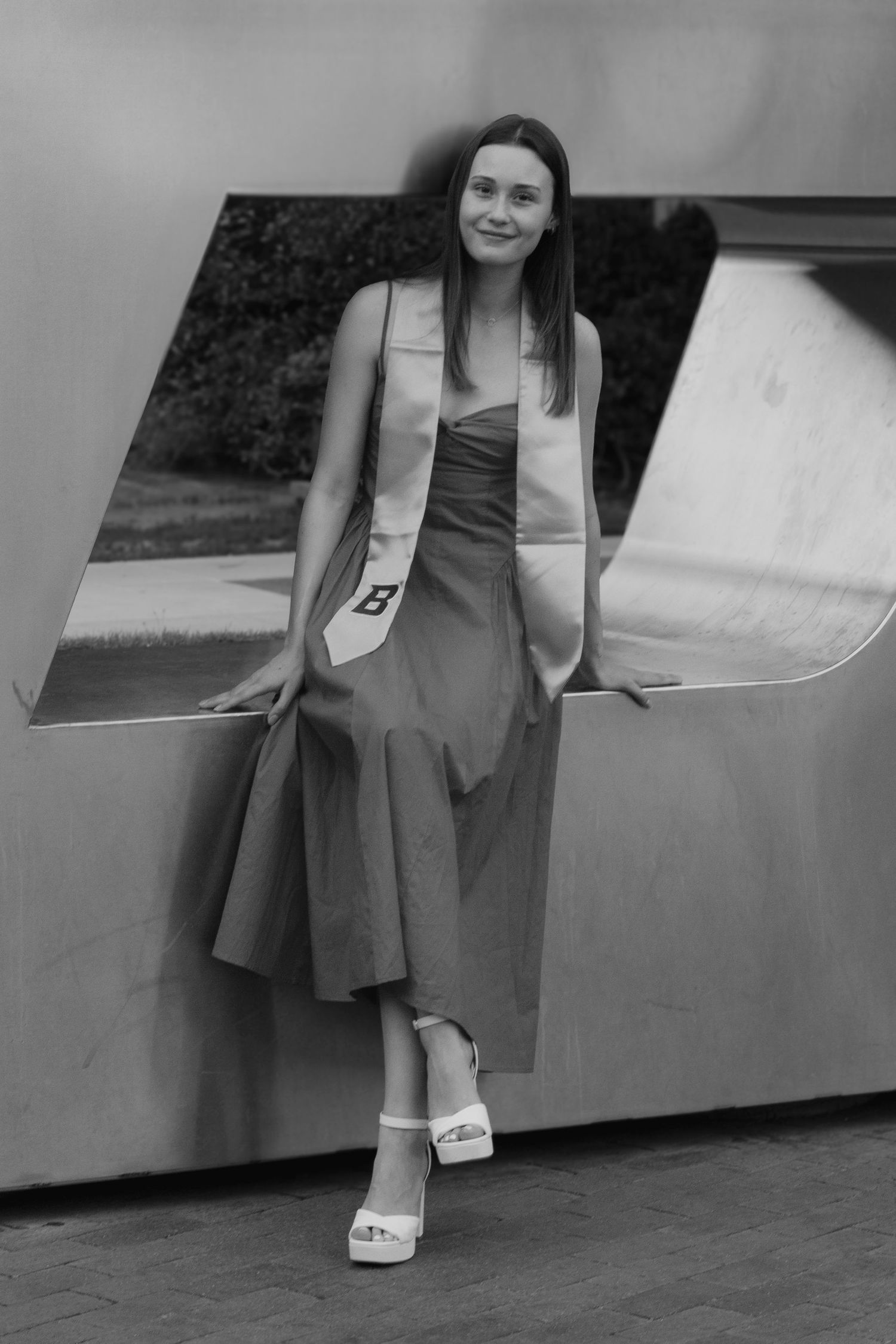 Black and white portrait of a graduate in a midi dress and stole leaning against a sculpture opening, wearing platform sandals.