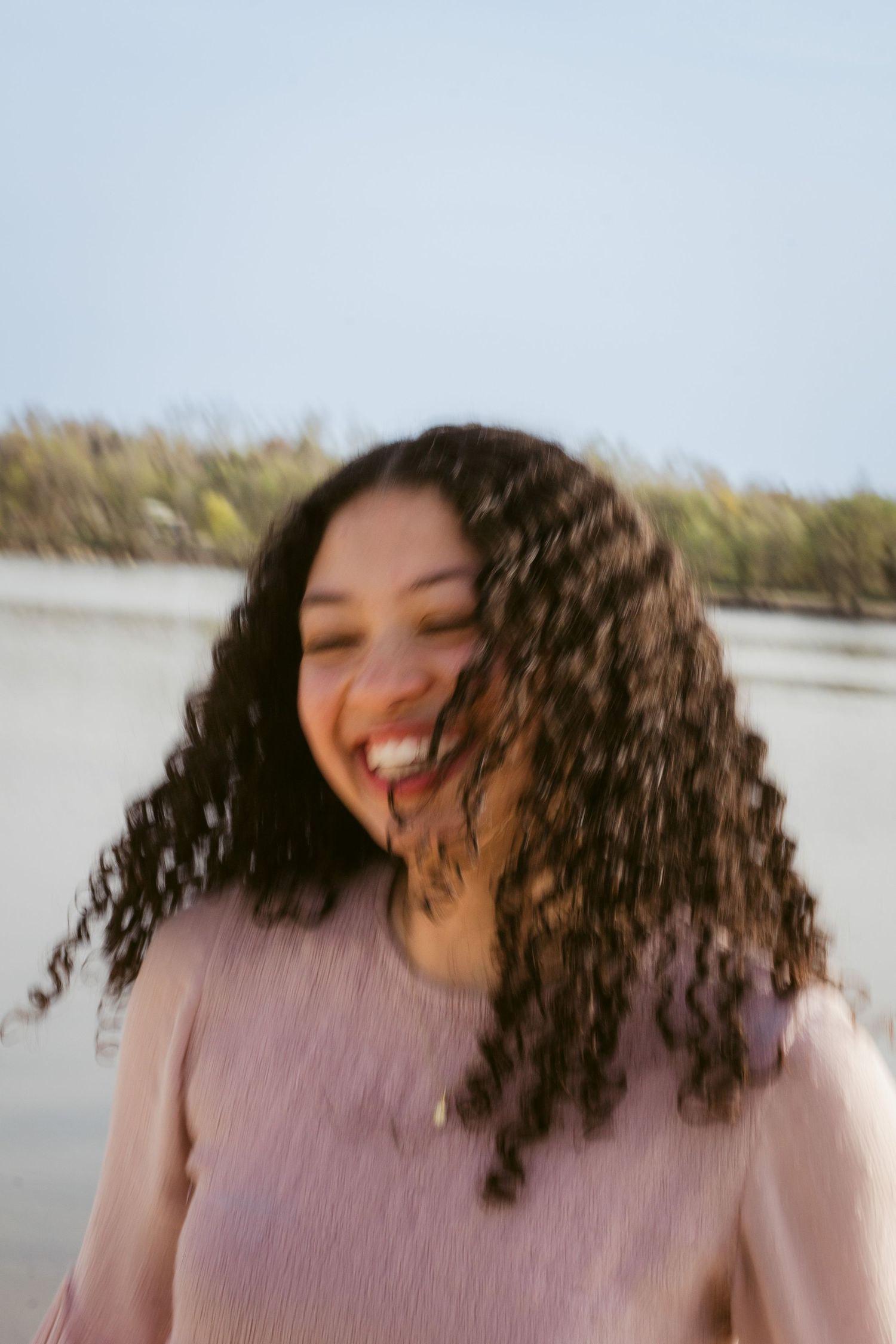 A joyful woman with long curly hair laughs in a motion-blurred portrait in a pink top beside a lake with trees behind her.