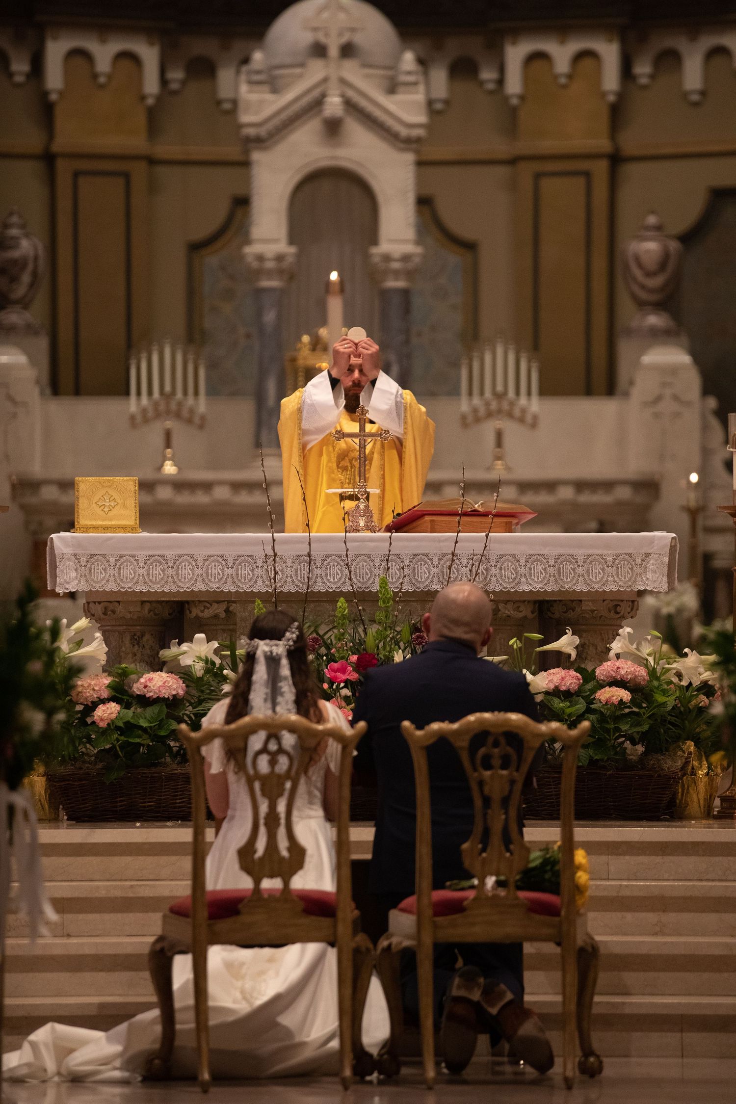 A bride and groom kneel before a church altar with flowers while a priest in gold vestments conducts the wedding Mass.