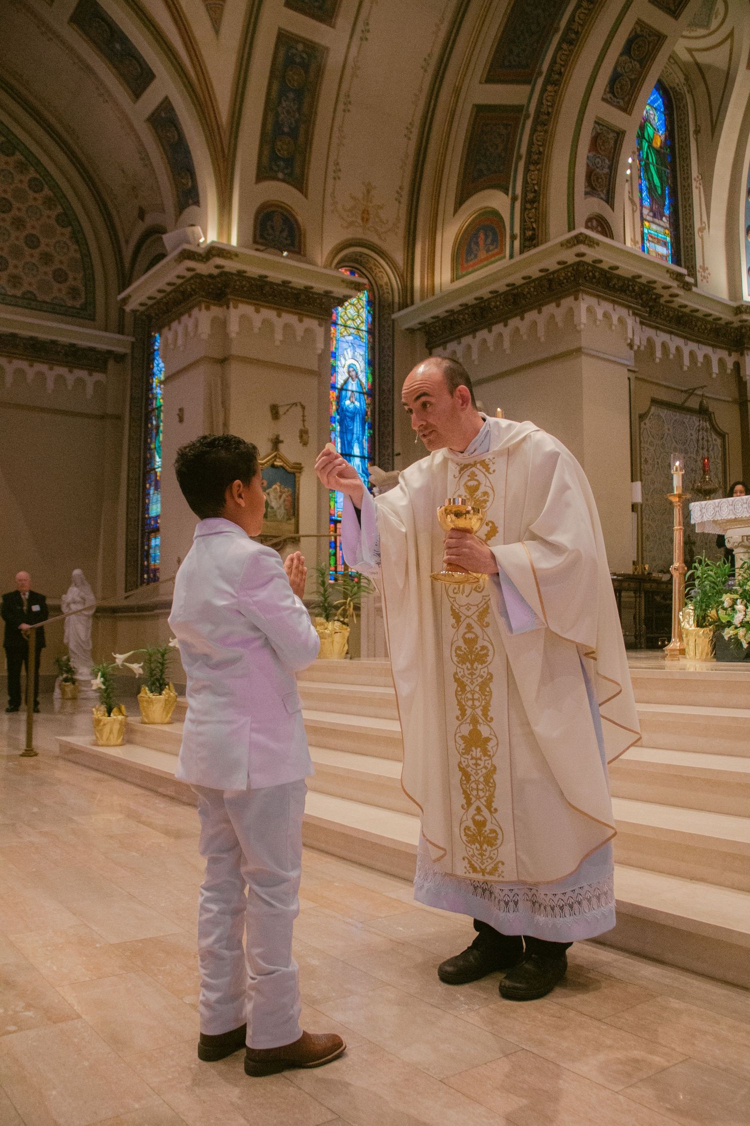A priest in white and gold vestments holds a golden chalice before a boy in a white suit at Catholic First Communion.