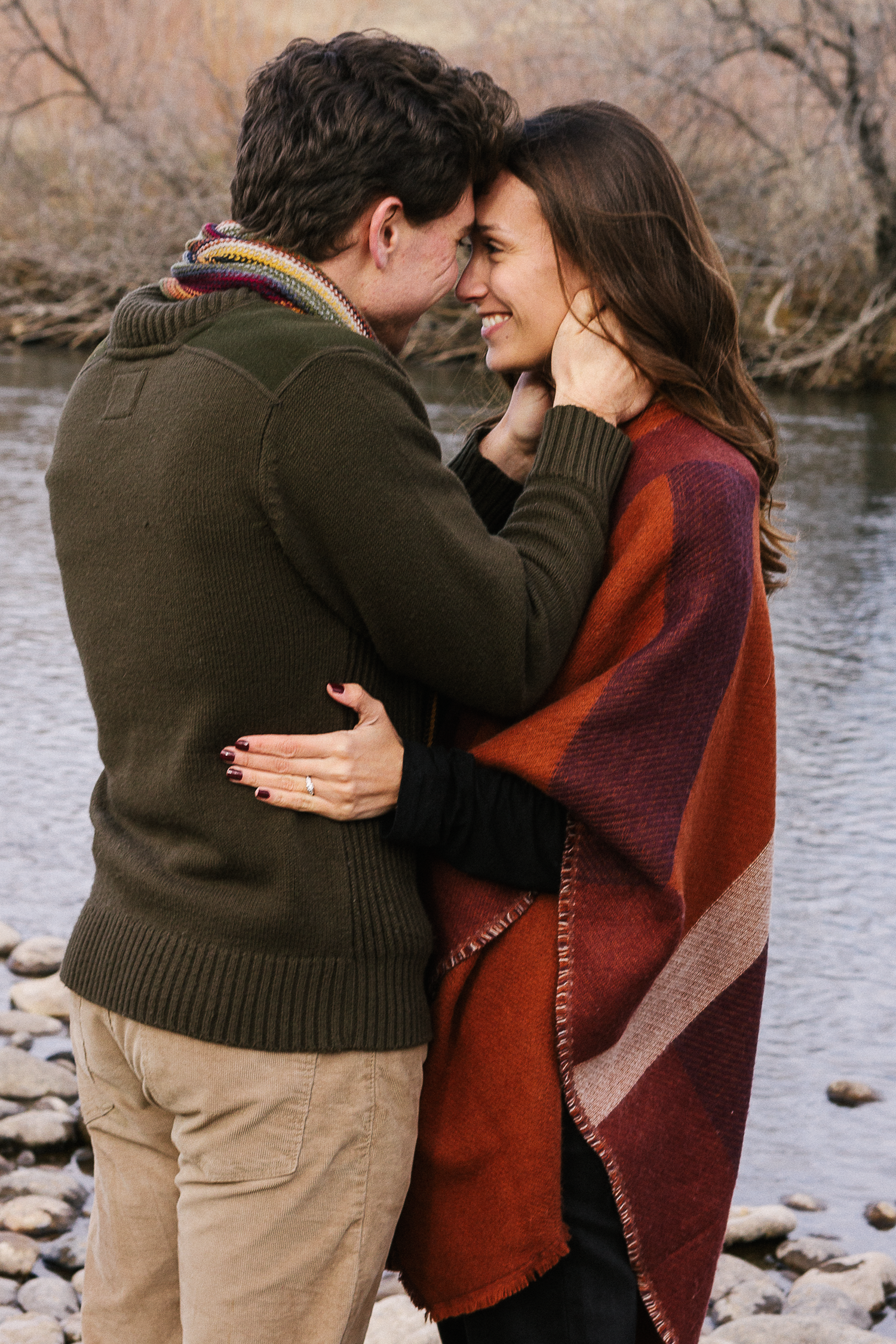 Couple touches foreheads in a warm embrace beside a river, she in a rust wrap and he in a green sweater.