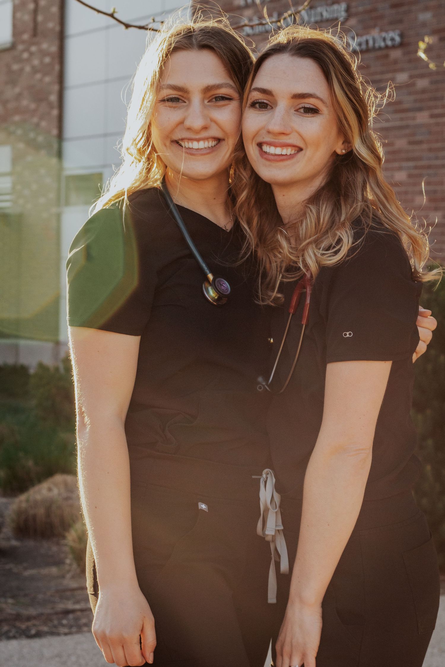 Two smiling nursing students in black scrubs with stethoscopes hug outside a brick campus building in warm golden sunlight.