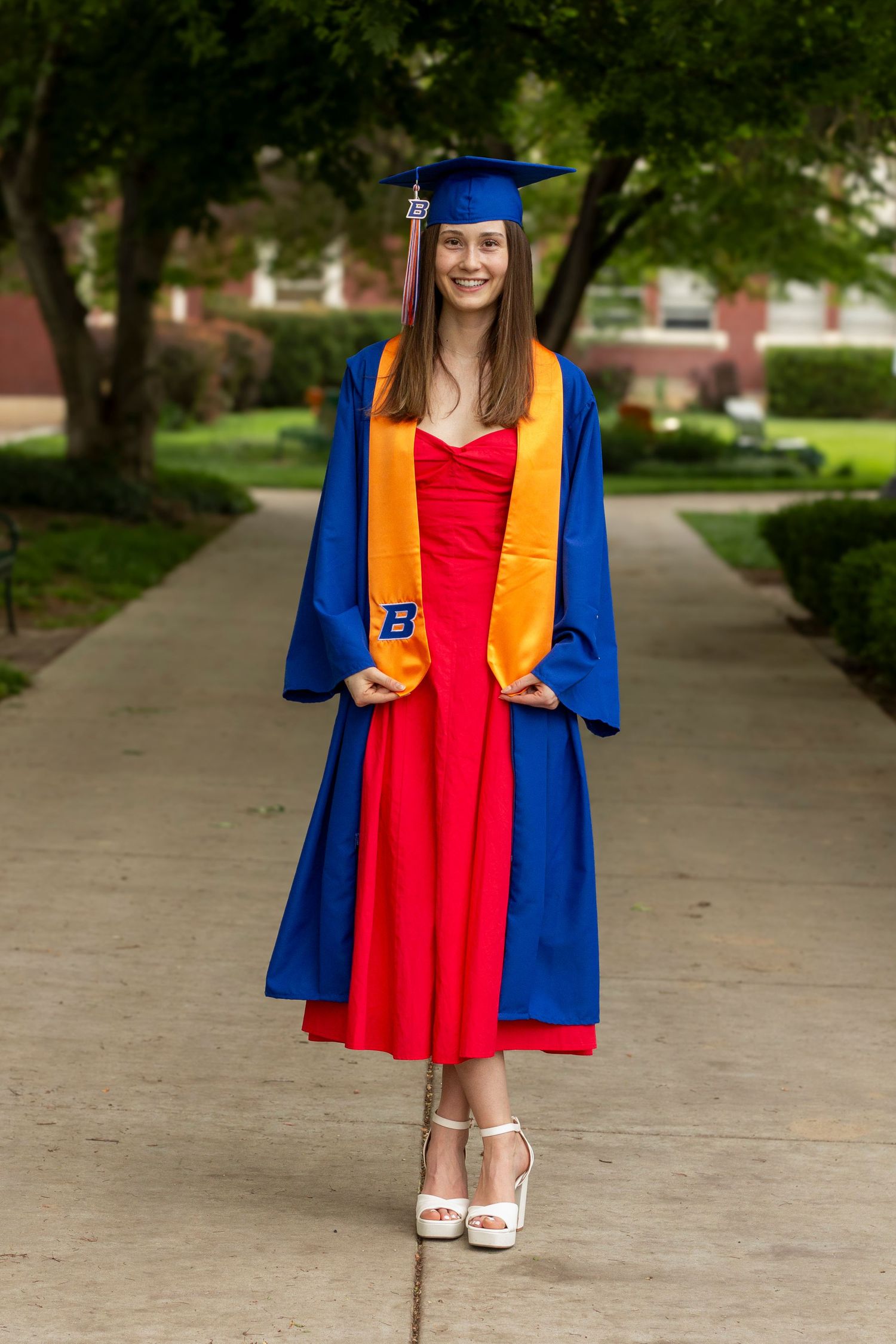 A smiling graduate in a blue gown, cap, and orange stole stands on a tree-lined brick campus walkway in a red dress.