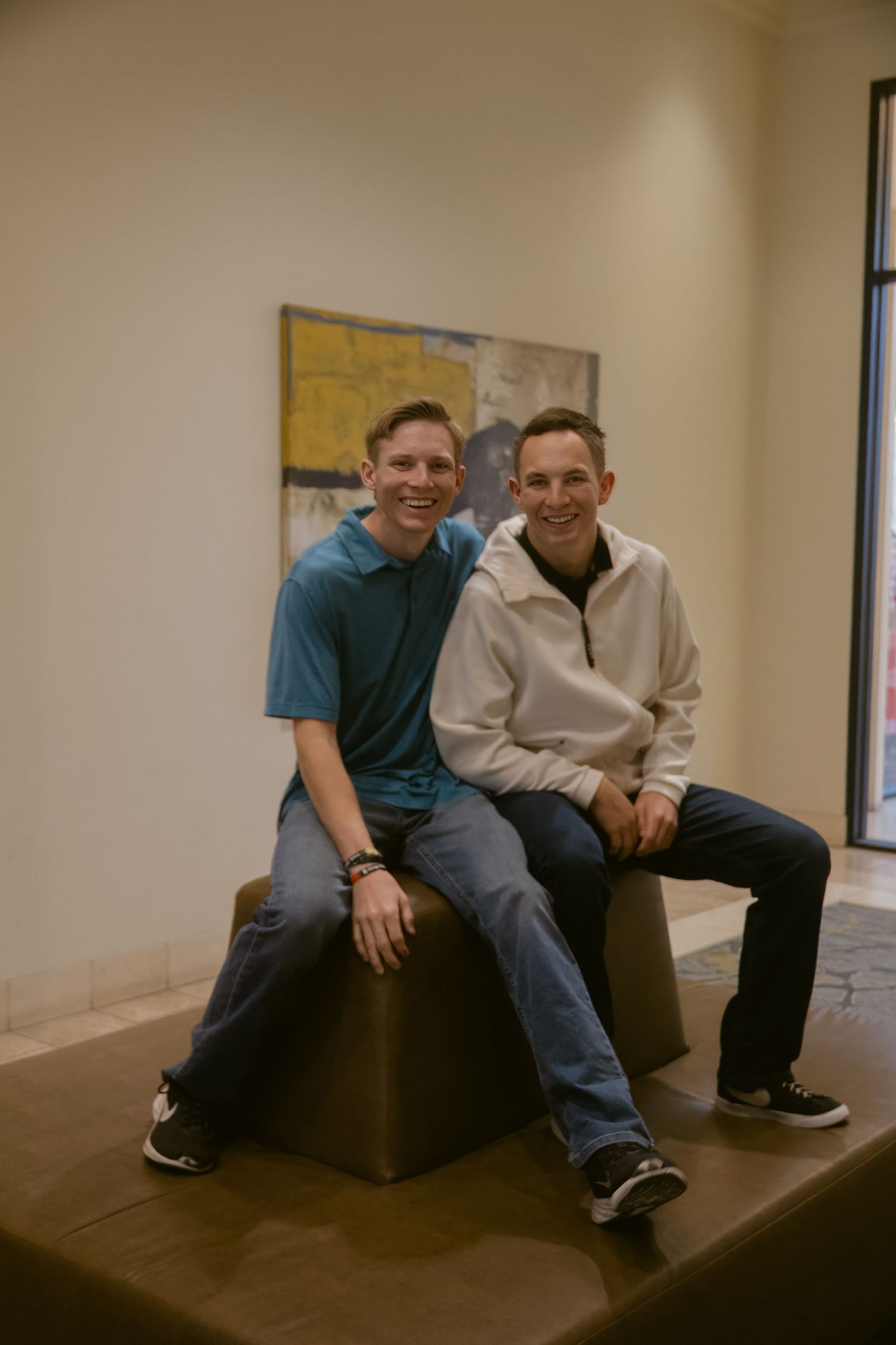Two smiling young men in casual clothes sit side by side on a leather ottoman in front of abstract wall art.