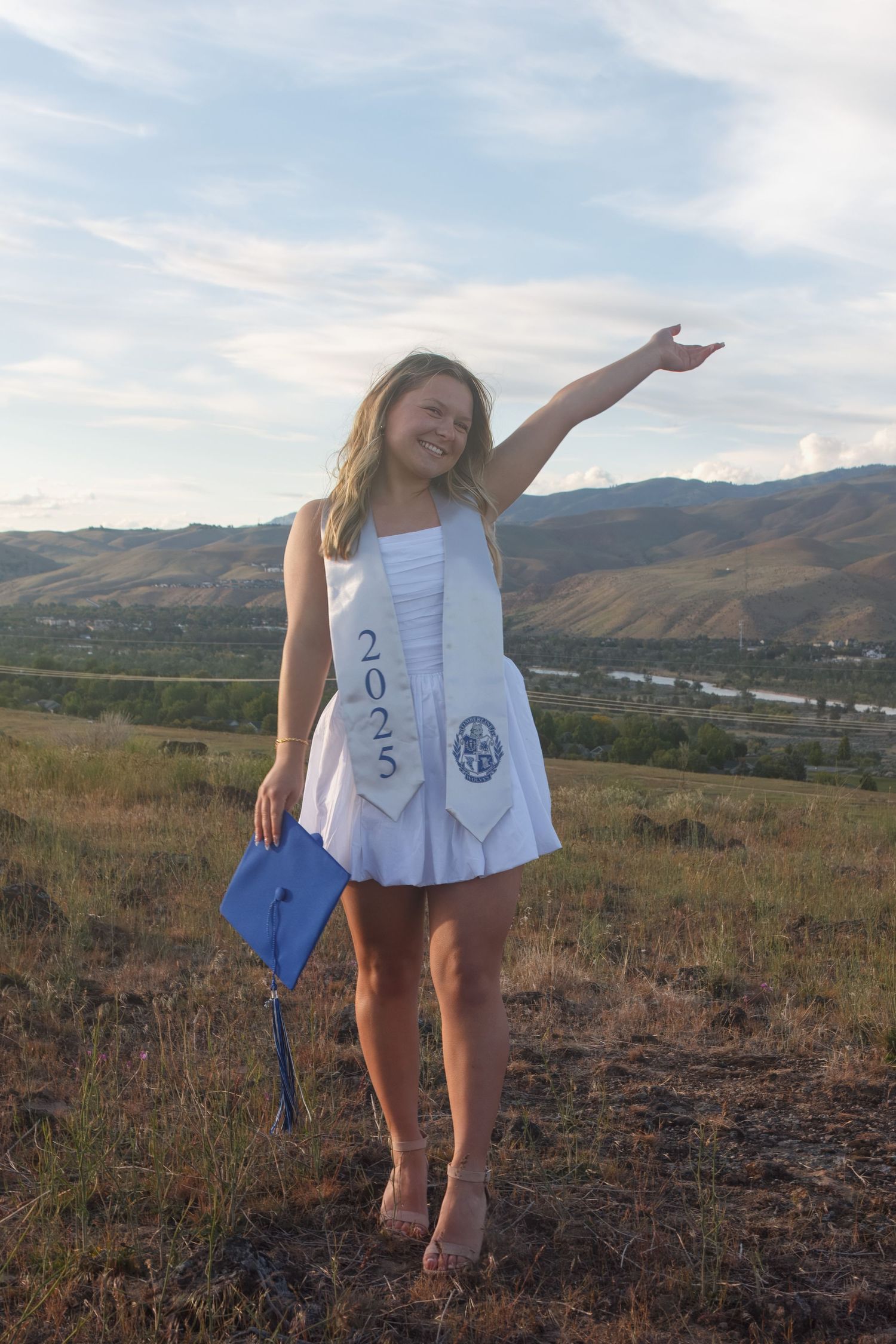 Smiling graduate in a white dress and 2025 stole holds a blue cap and raises one arm on a hillside overlooking a valley and river.
