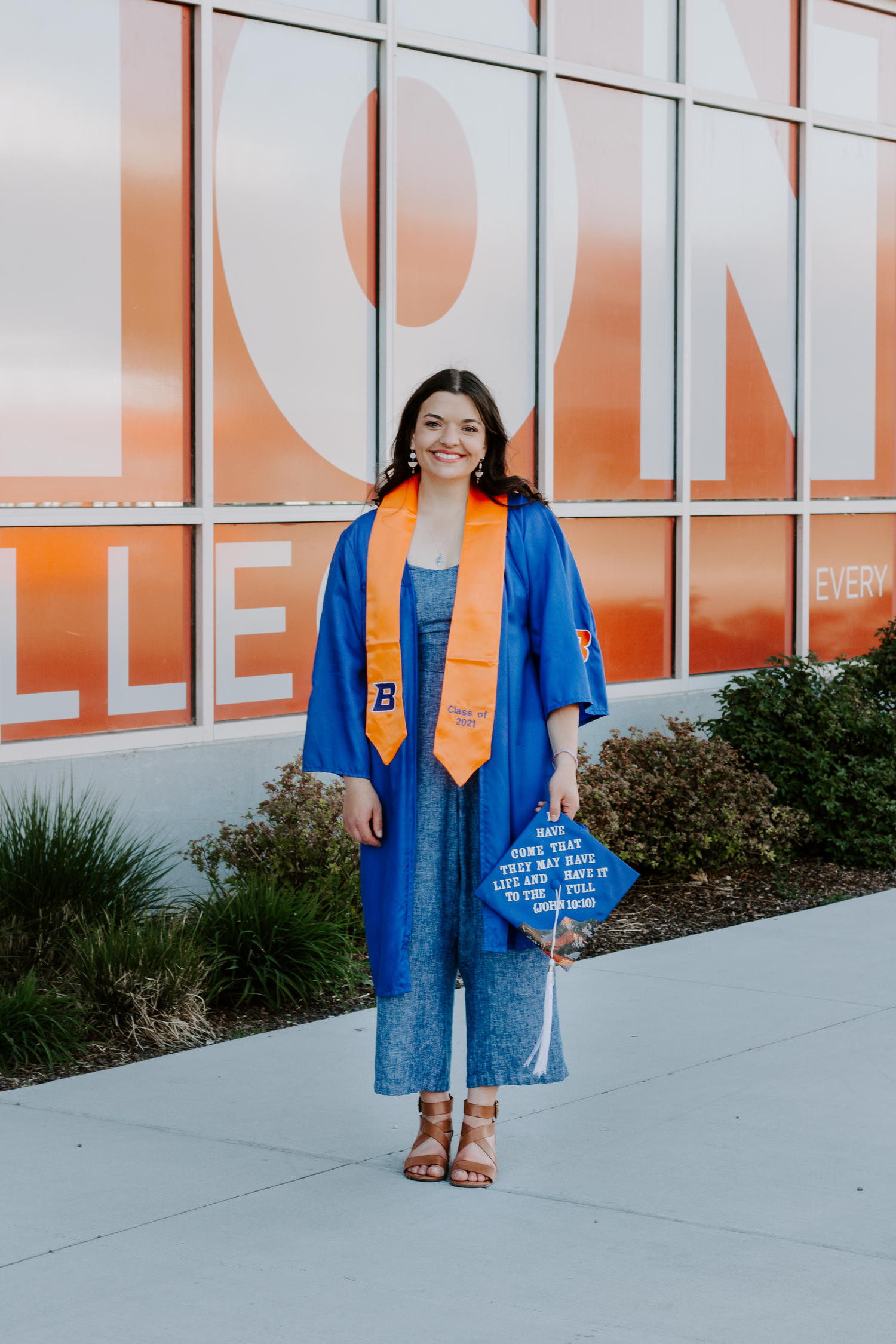 Smiling graduate in a blue gown with orange stole holds a decorated graduation cap while posing outside a building with orange windows.