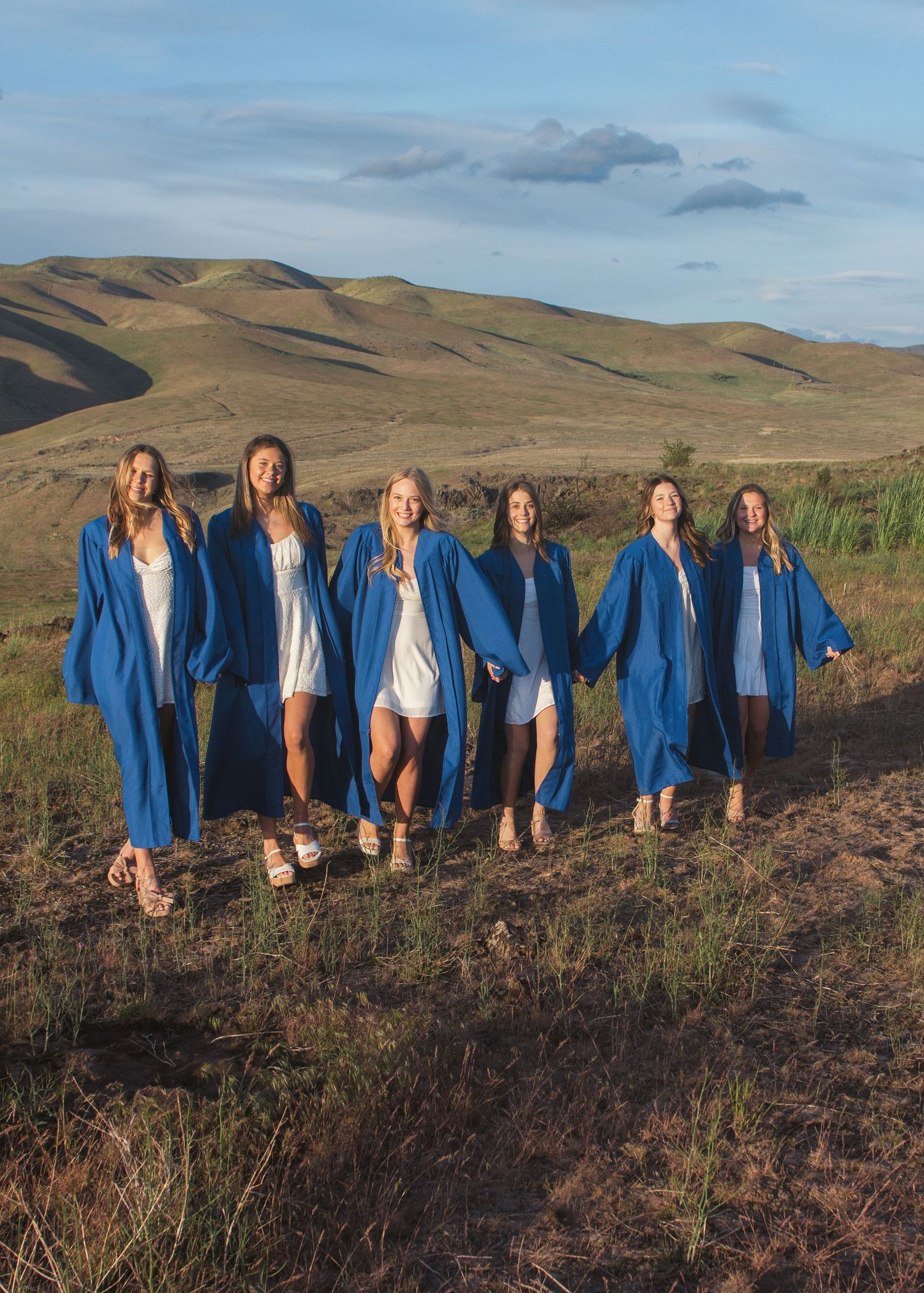Six graduates in blue gowns and white dresses walk arm in arm across a grassy hillside under a soft blue sky.