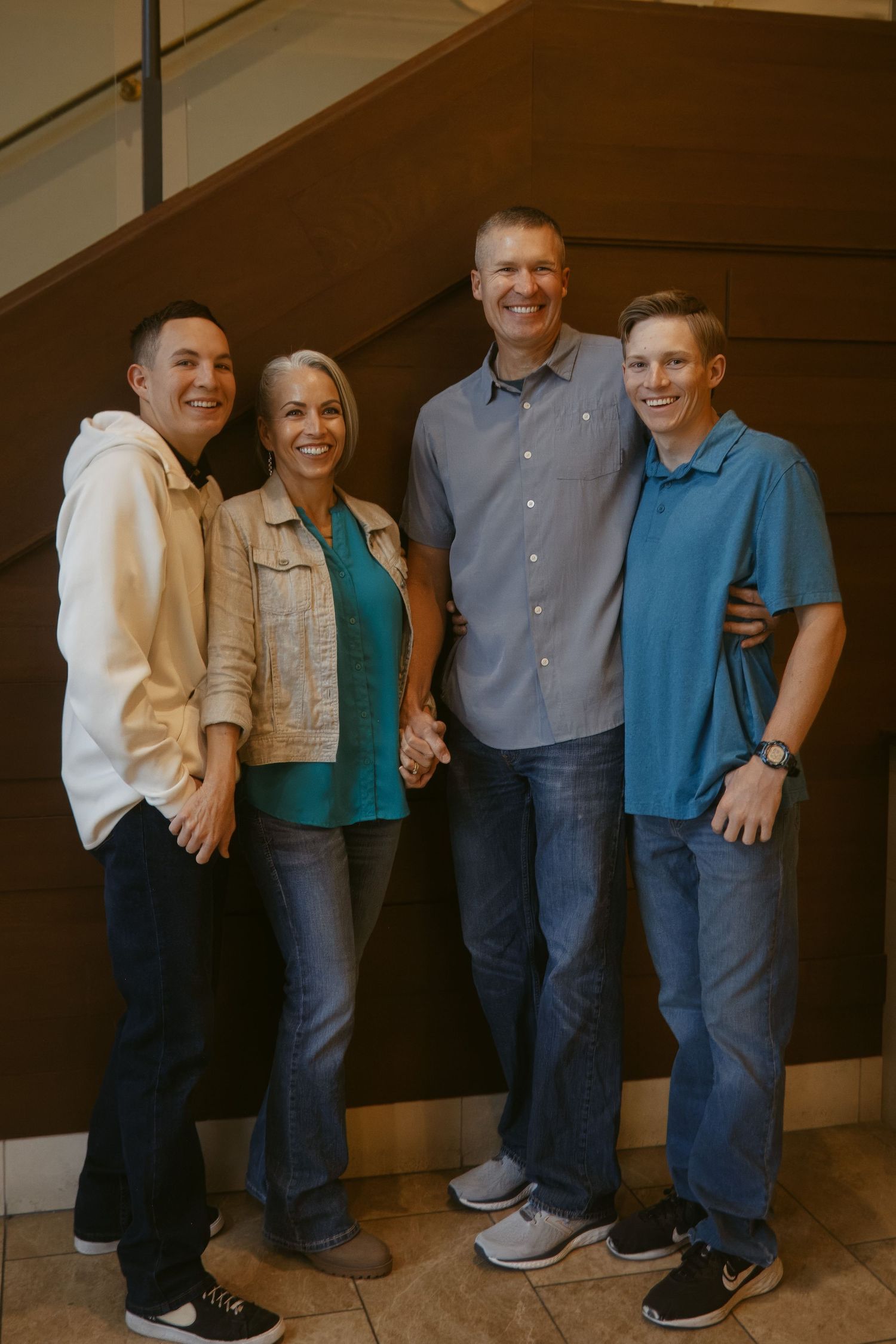 Four smiling family members, a woman and three men in casual clothes, pose together in front of a wood-paneled staircase.