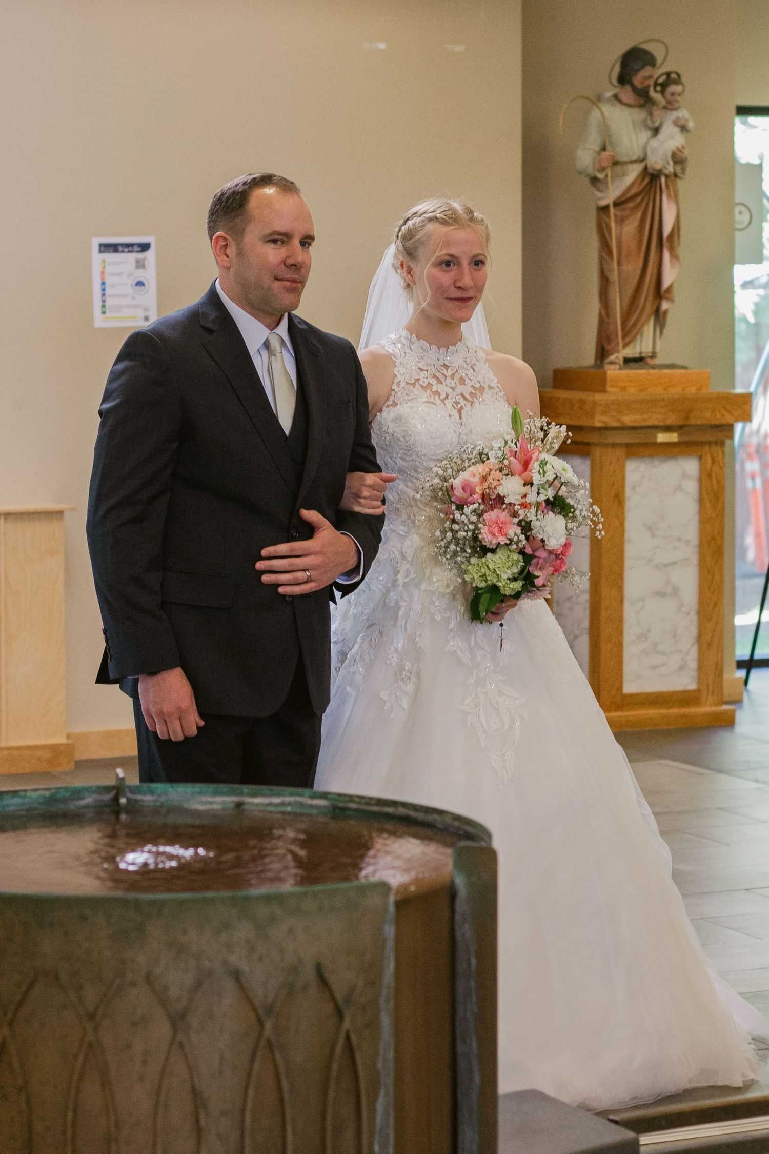 A groom in a dark suit escorts a bride in a lace wedding gown holding a pink bouquet past a baptismal font in a catholic church.
