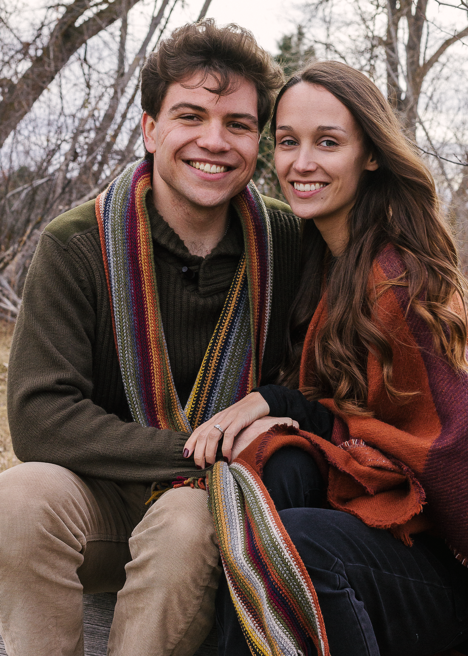 Smiling couple in cozy fall sweaters and a striped scarf sit close together outdoors among bare trees.