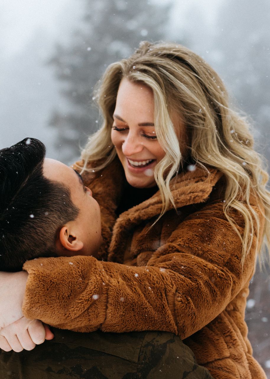 A smiling blonde woman in a brown faux fur coat embraces a dark-haired man as snow falls around them.