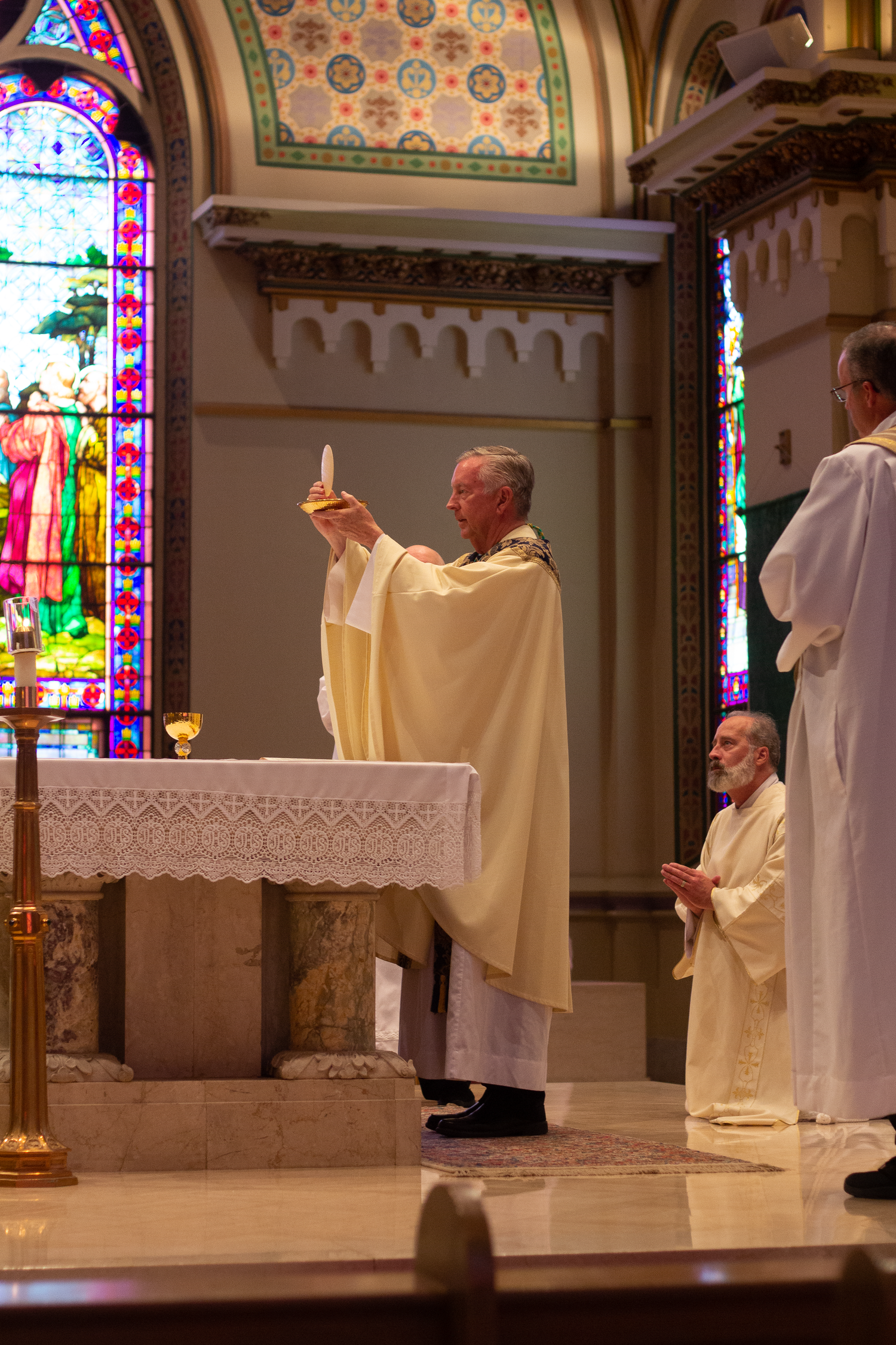 A priest in cream vestments raises the Eucharist at the altar during Mass beneath stained glass windows in a Catholic church.