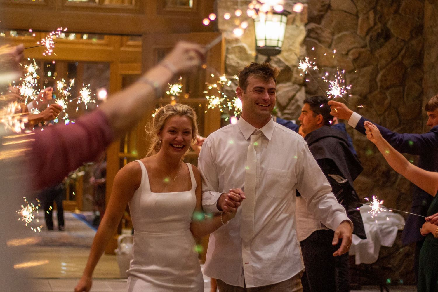 A smiling bride and groom walk hand in hand through a sparkler send-off as guests hold up glowing sparklers at dusk.